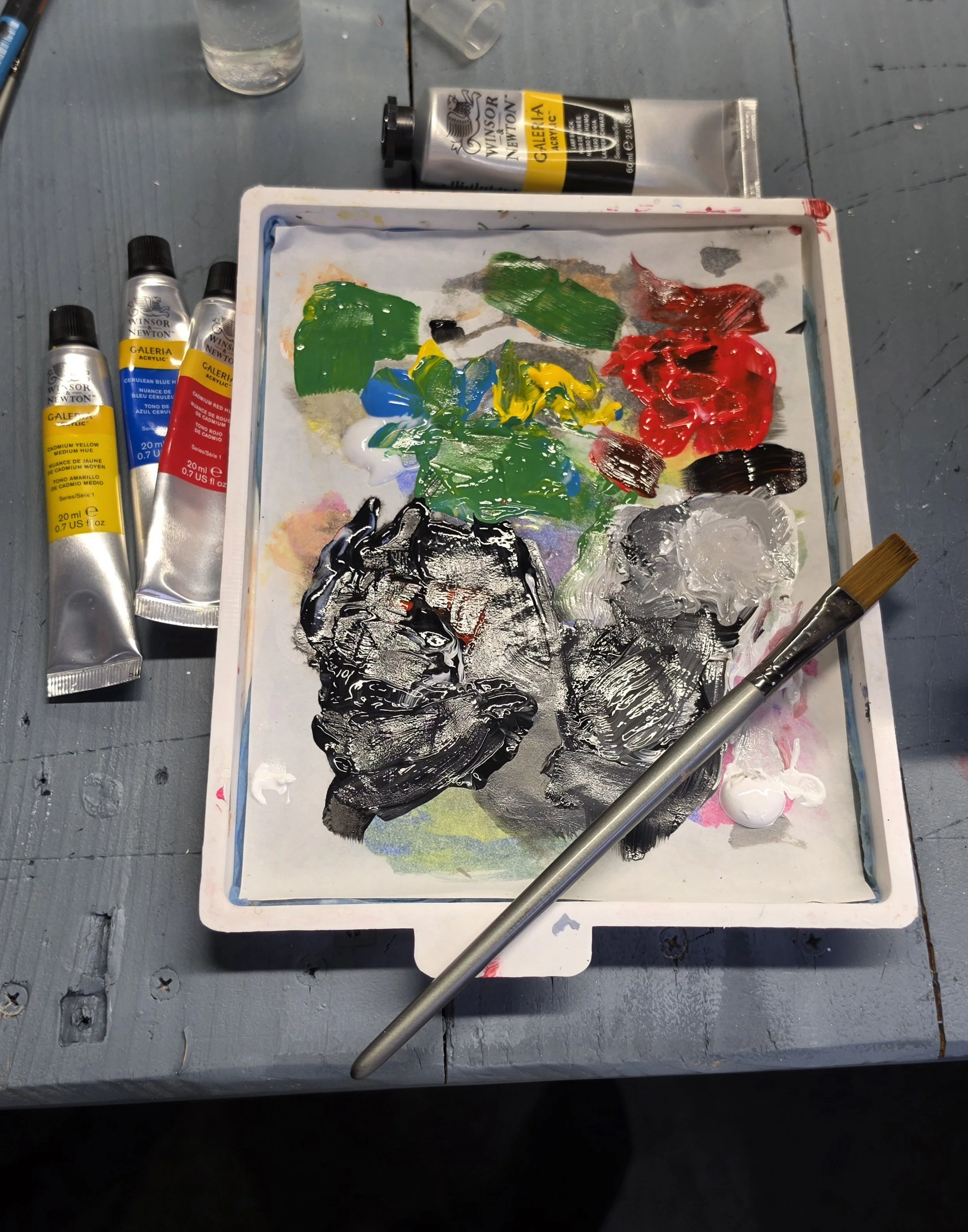 A palette with various blobs of paints including green, blue, yellow, red, black, and white, with tubes of Winsor & Newton acrylic paint nearby on a wooden surface.