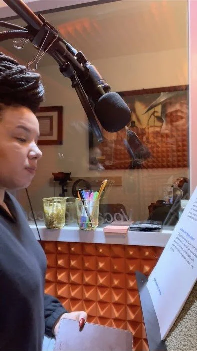 Woman standing in front of a microphone, reading from a sheet of paper in a recording studio.