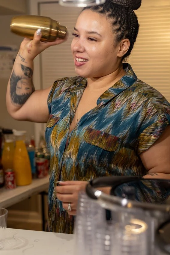 Woman with tattoos and braided hair smiling while holding a brass cocktail shaker in her right hand