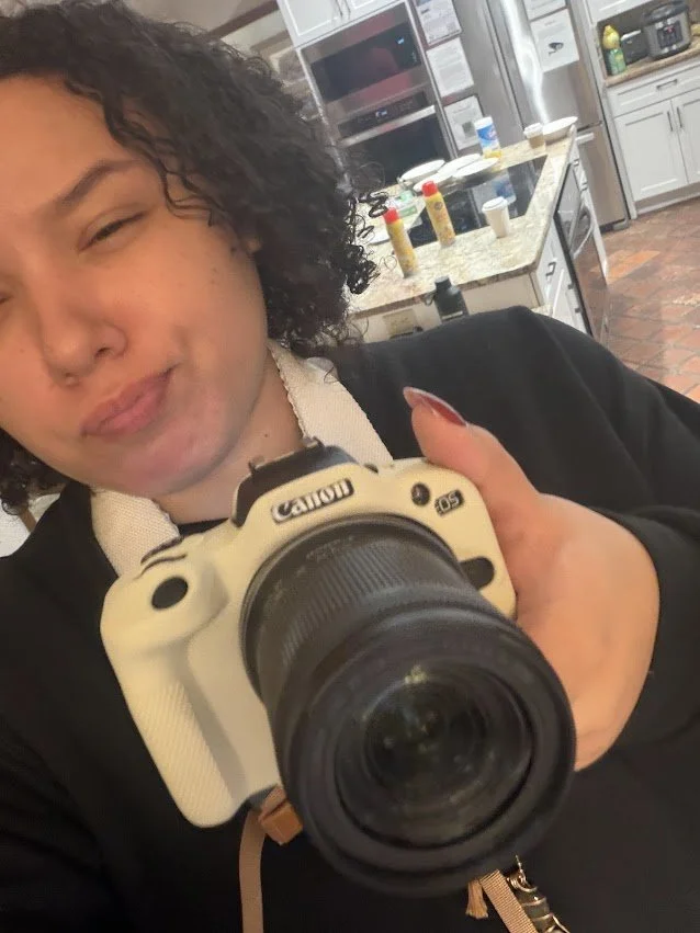 Young woman taking a selfie with a professional camera, in a kitchen with various condiments on the counter in the background.