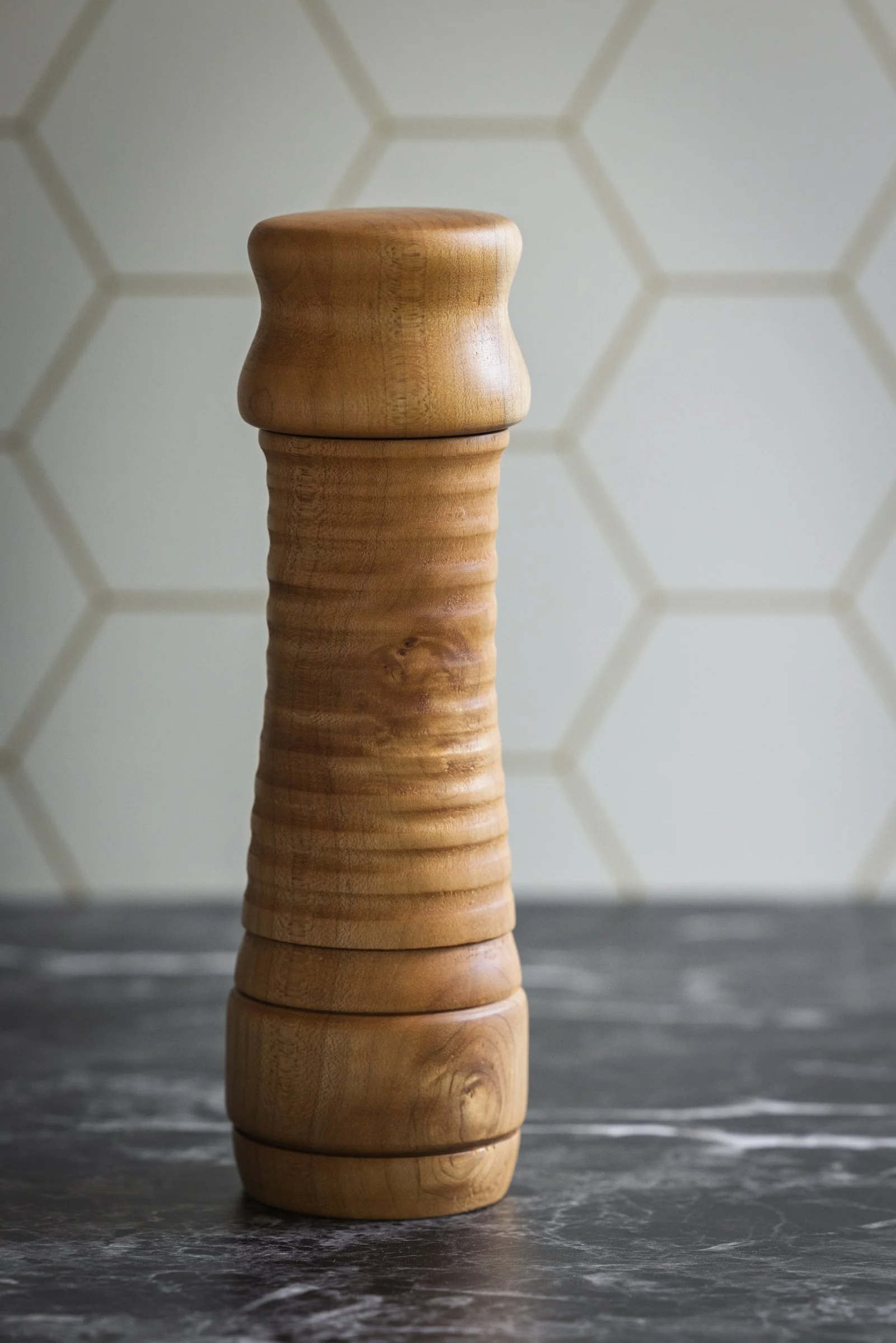 A pepper or salt ginder made from maple sitting on a dark granite countertop with a tiled wall in the background.