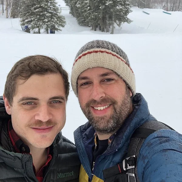 Two men smiling outdoors in a snowy landscape, wearing winter jackets and a knit hat, with snow-covered trees in the background.