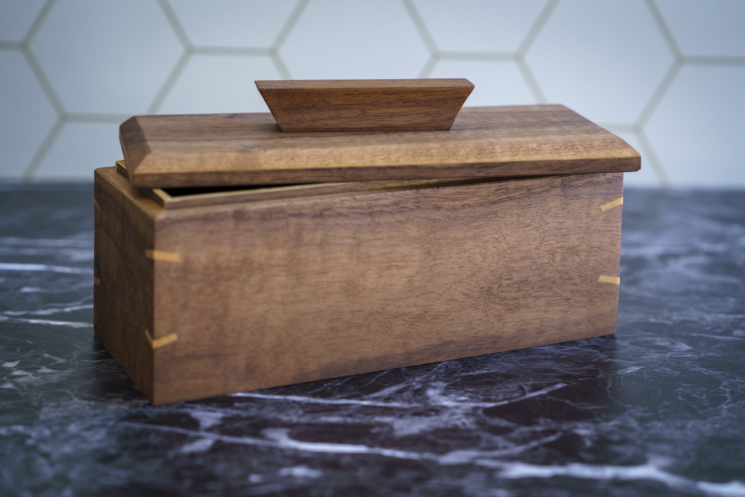A wooden box with an askew lid, placed on a dark granite surface. The background has a light-colored wall with a hexagonal pattern.