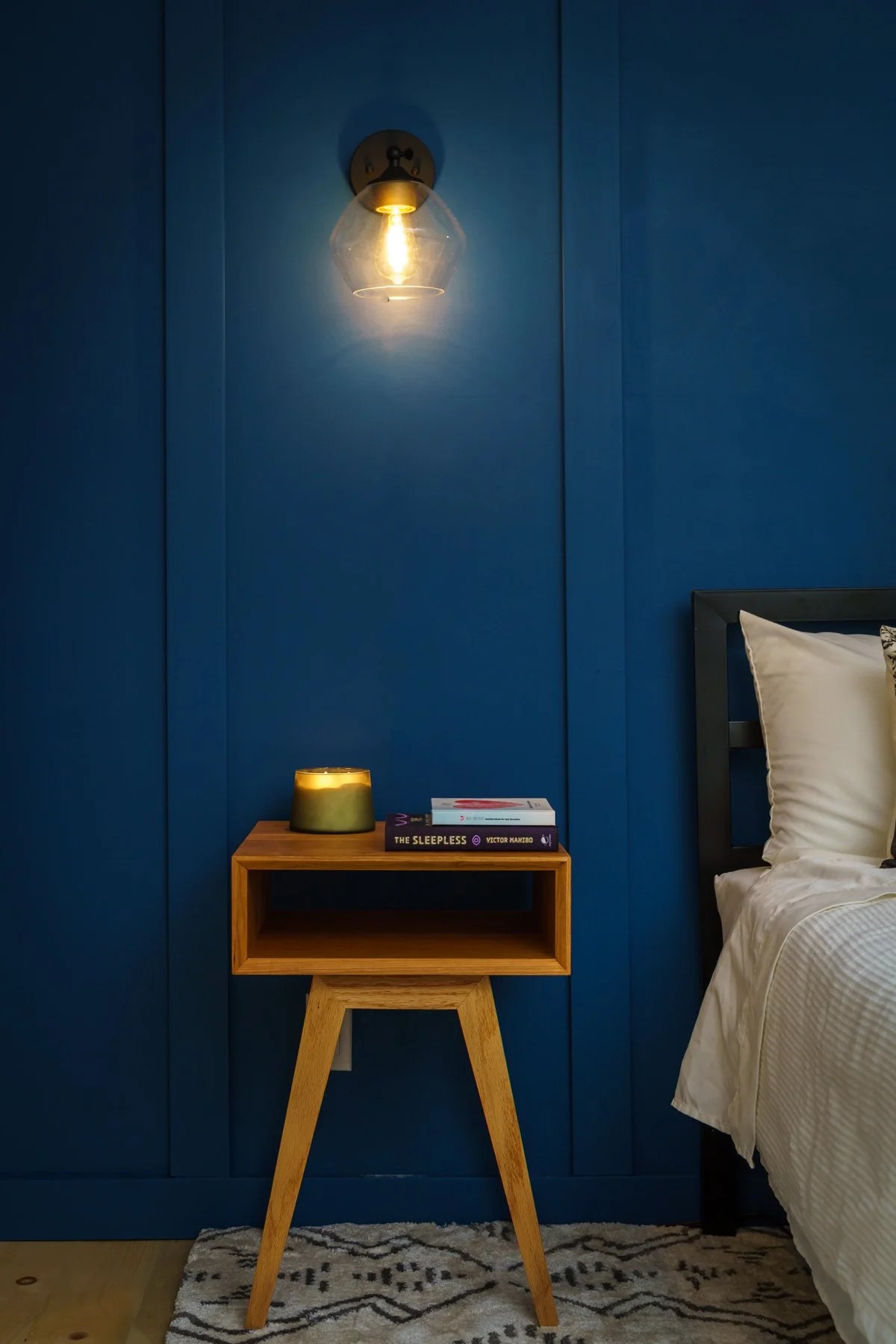 A bedroom with a blue wall, a wall-mounted light fixture, a wooden nightstand with a hardcover book, a small greenish lamp, and part of a bed with white bedding and a black bed frame.