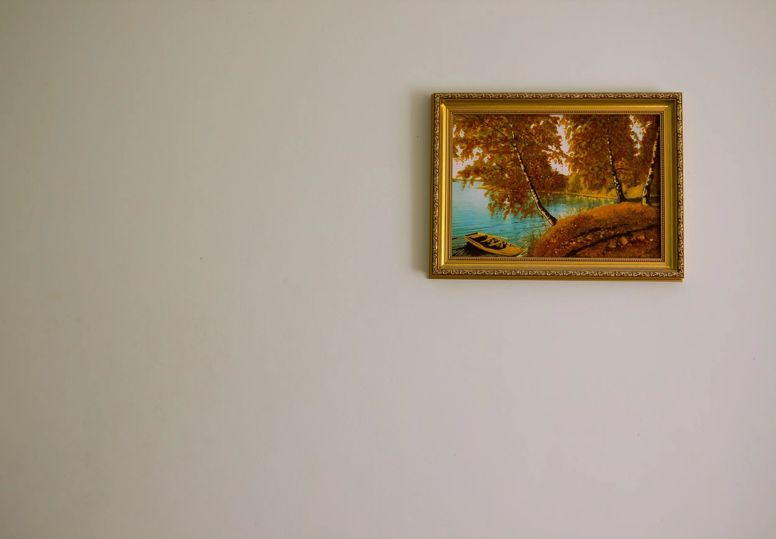 A framed painting of a lakeside scene with trees, a boat, and calm water, hung on a plain white wall.
