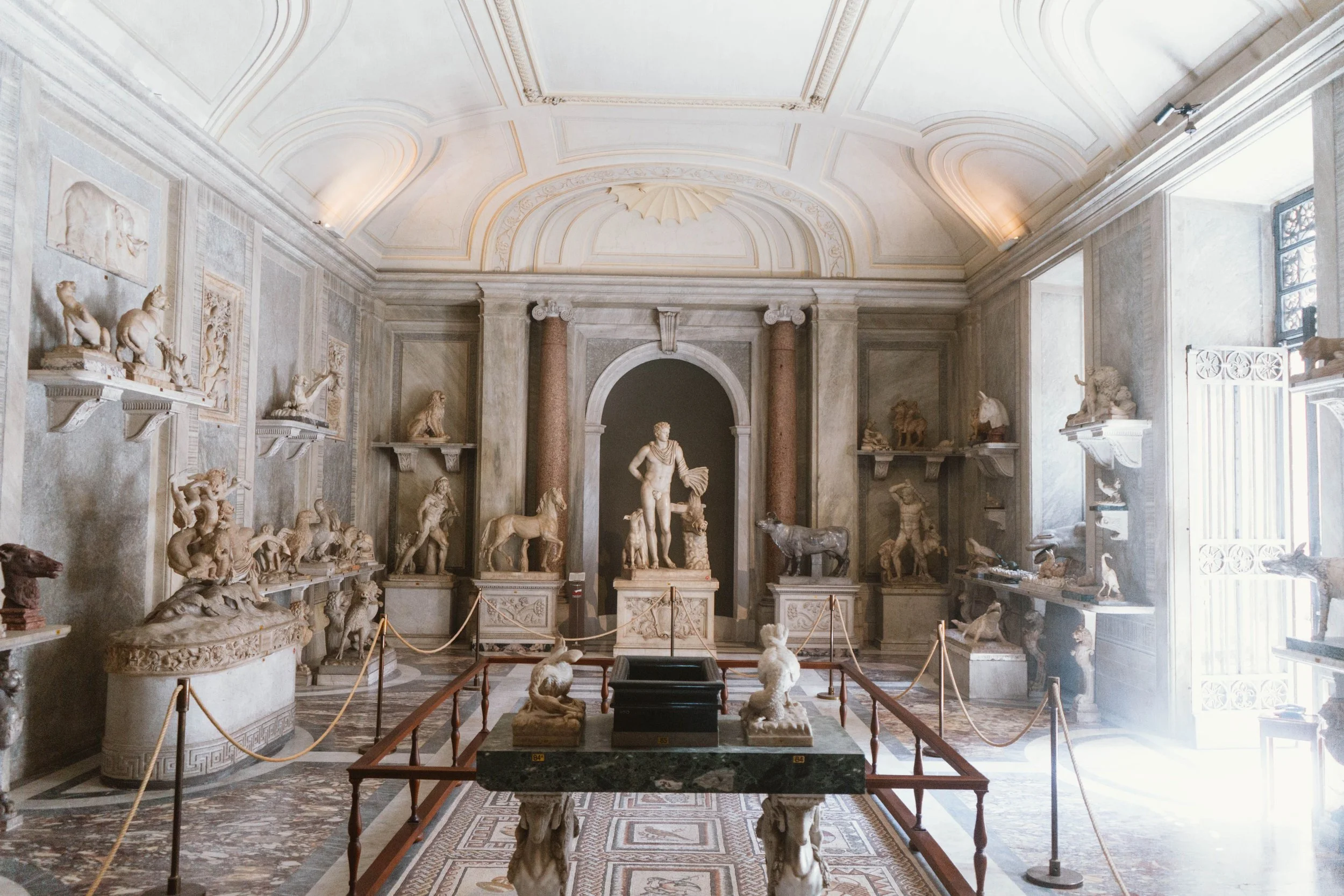 A museum room with classical sculptures, mostly Greek or Roman style, displayed on shelves and pedestals. The room features ornate ceiling decorations, columns, and large windows allowing natural light to illuminate the sculptures.