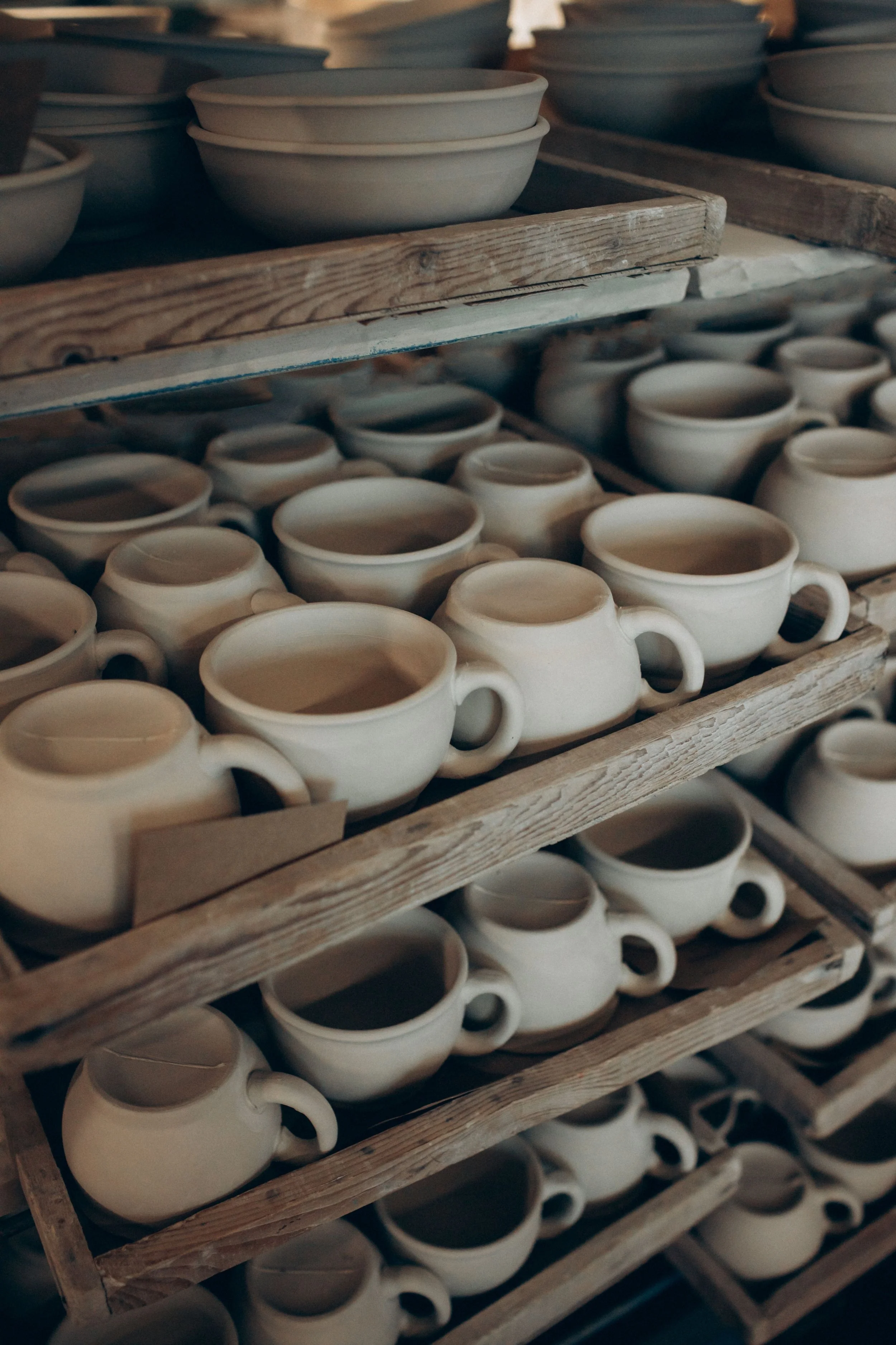 Multiple unfinished pottery bowls with handles are stacked on wooden shelves in a pottery studio.