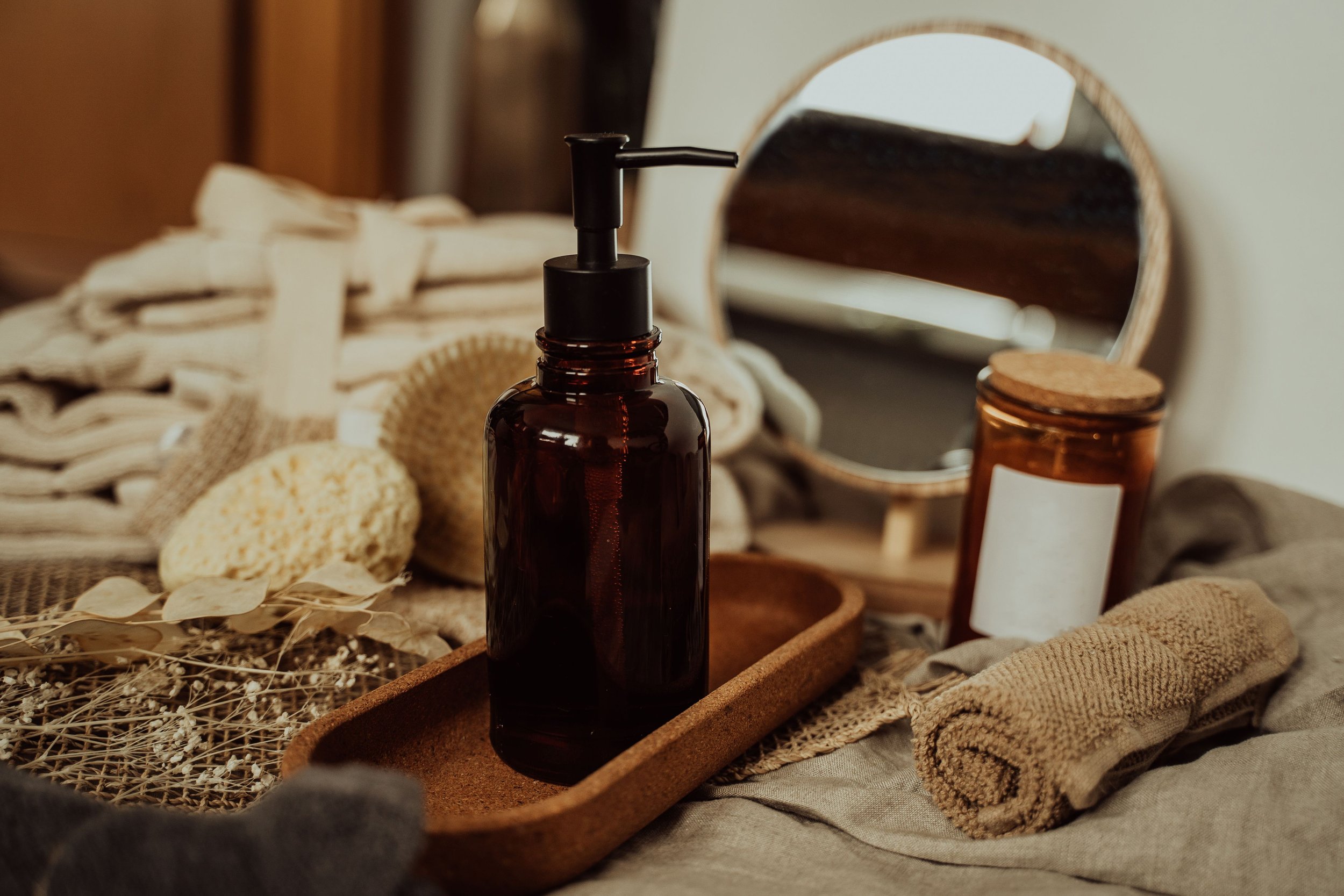 A brown glass bottle with a pump dispenser on a wooden tray surrounded by natural and skincare items, including a mirror, a corked bottle, towels, and dried botanical elements.