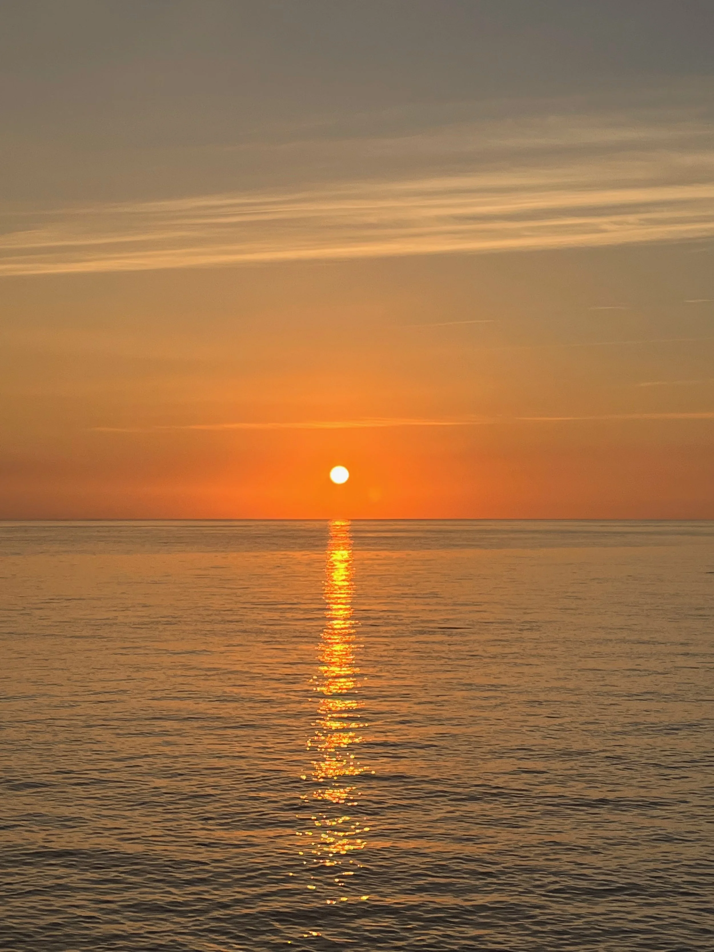 A sunset over the ocean with the sun near the horizon, reflecting a golden trail on the water. The sky has soft clouds with a warm glow, creating a peaceful and scenic view.
