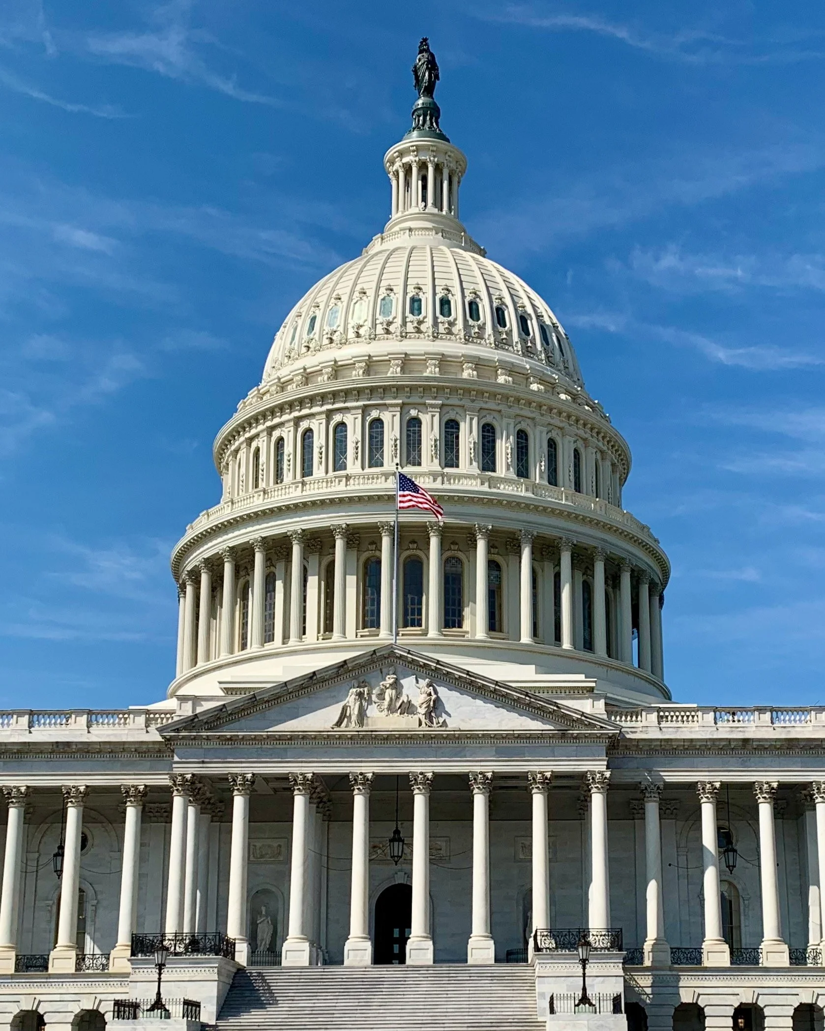 The United States Capitol building with a large dome, columns, stairs, and an American flag in front, against a blue sky.