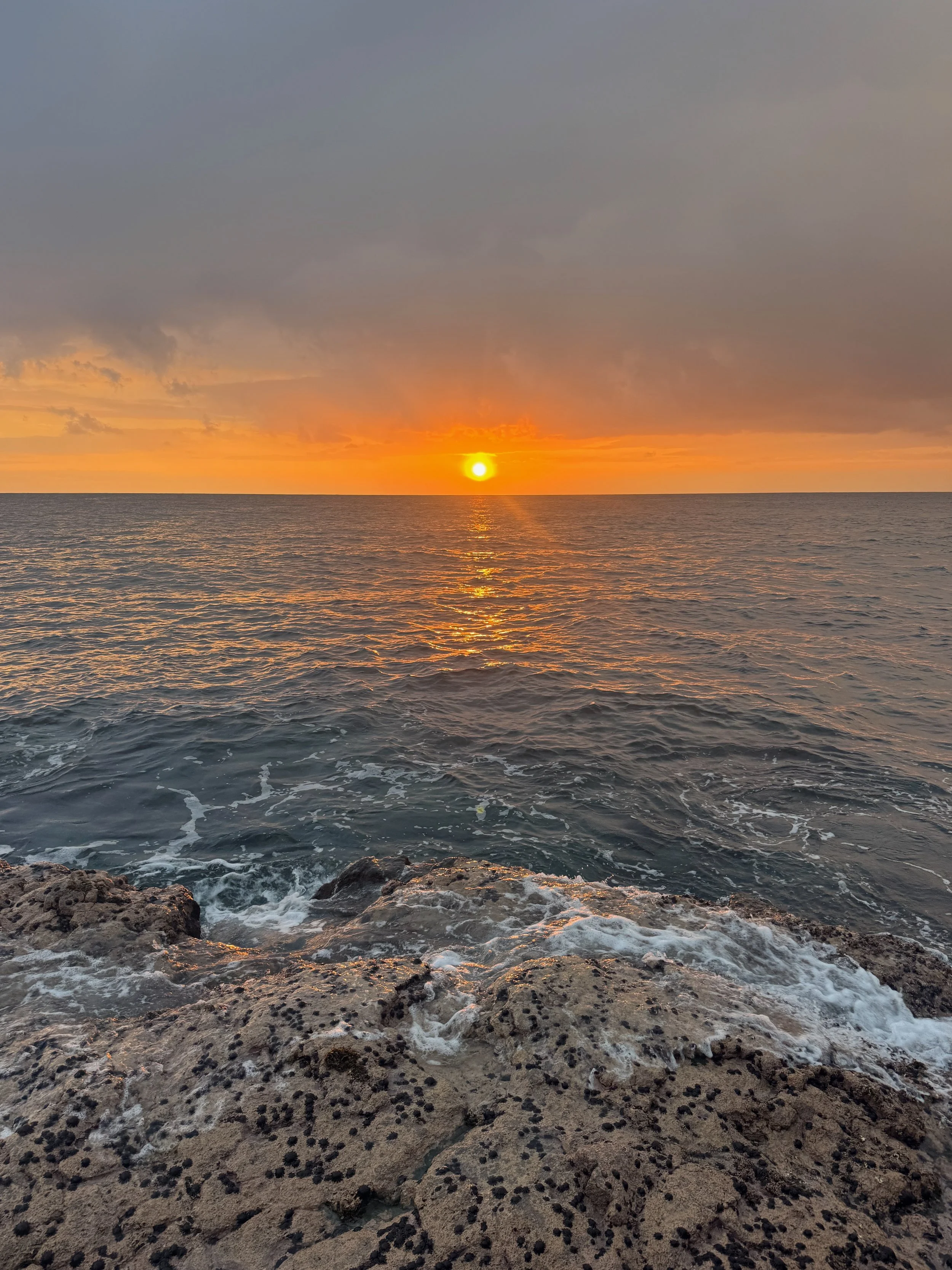 A sunset over the ocean with dark clouds in the sky, reflecting orange and yellow hues on the water, with rocky shoreline in the foreground.