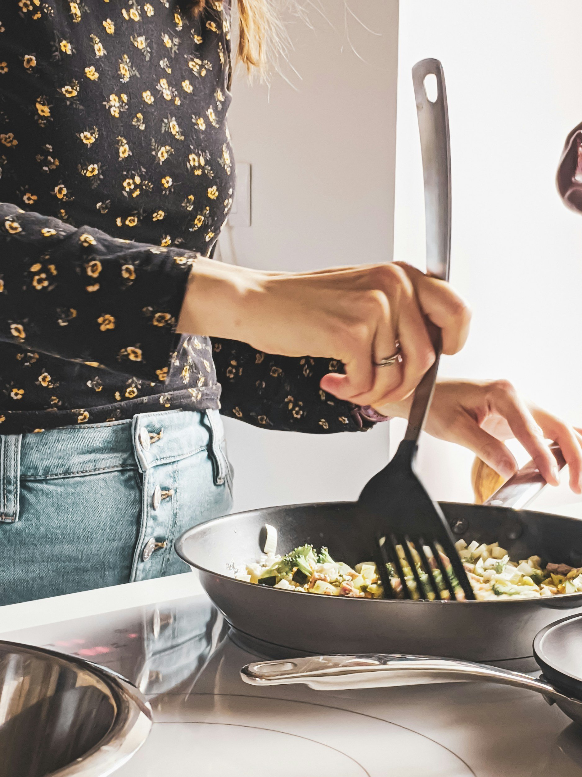 A person cooking pasta on an electric stovetop in a kitchen, wearing a black shirt with yellow floral patterns and blue jeans.