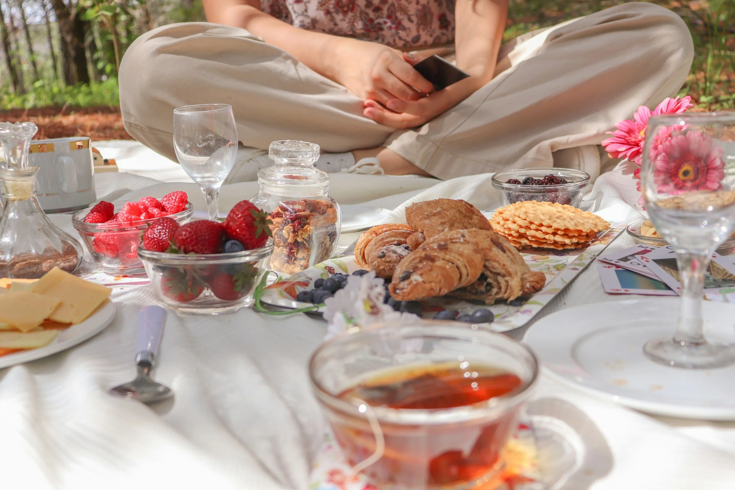 A person sitting cross-legged outdoors at a picnic table with a spread of various foods, including berries, pastries, cheese, and drinks, with trees in the background.
