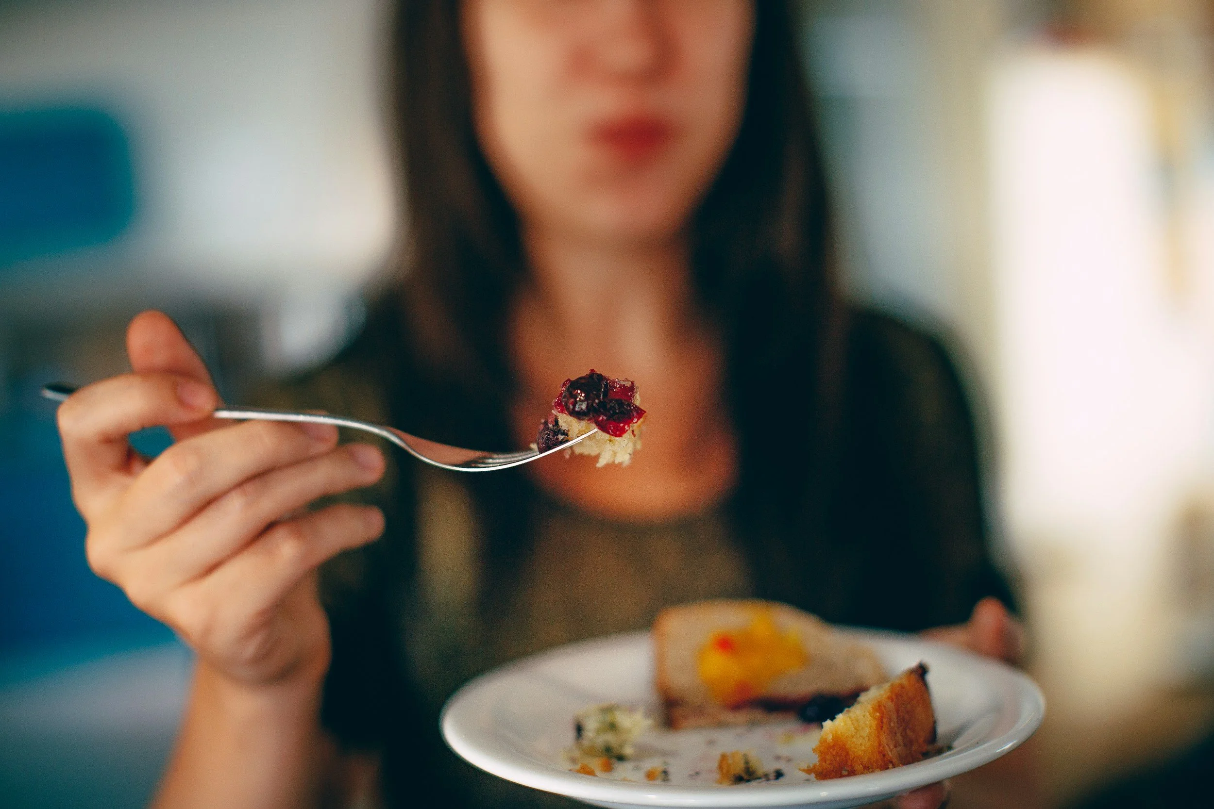 A woman holding a fork with a bite of berry-topped dessert in focus, while her face is blurred in the background. She is sitting at a table with a plate of cake and other desserts.