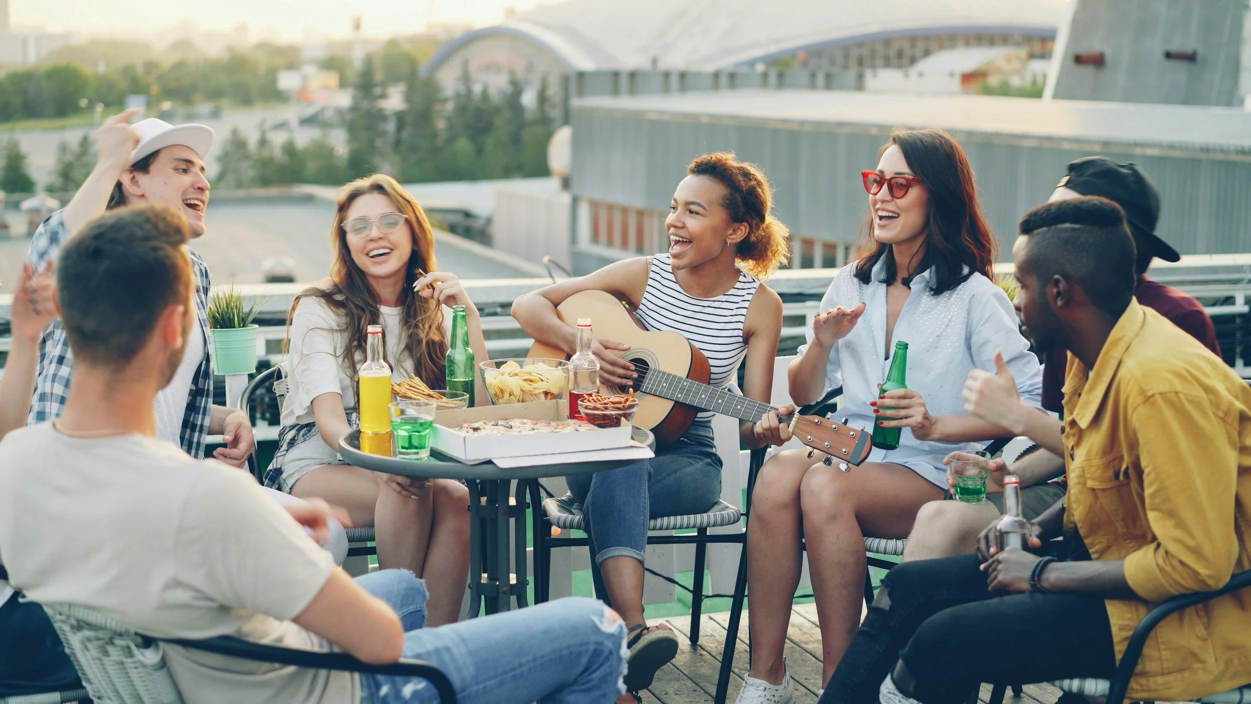 A diverse group of friends enjoying a rooftop party with food and drinks, one is playing guitar, all are laughing and having a good time.