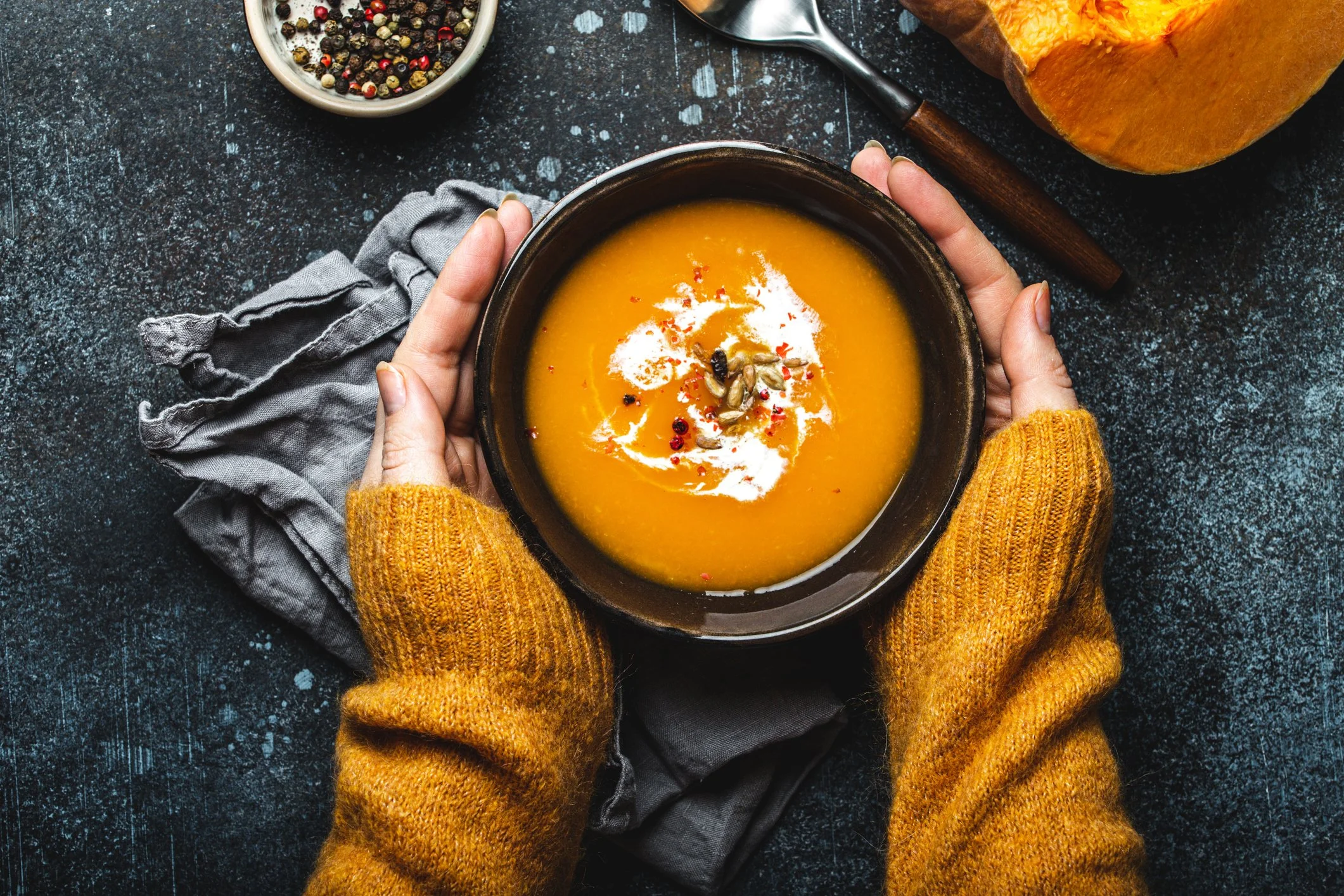 A person wearing yellow sweater holding a bowl of orange soup garnished with seeds and red pepper flakes, on a dark textured surface with a grey cloth, a bowl of mixed peppercorns, a silver spoon, and a piece of pumpkin in the background.