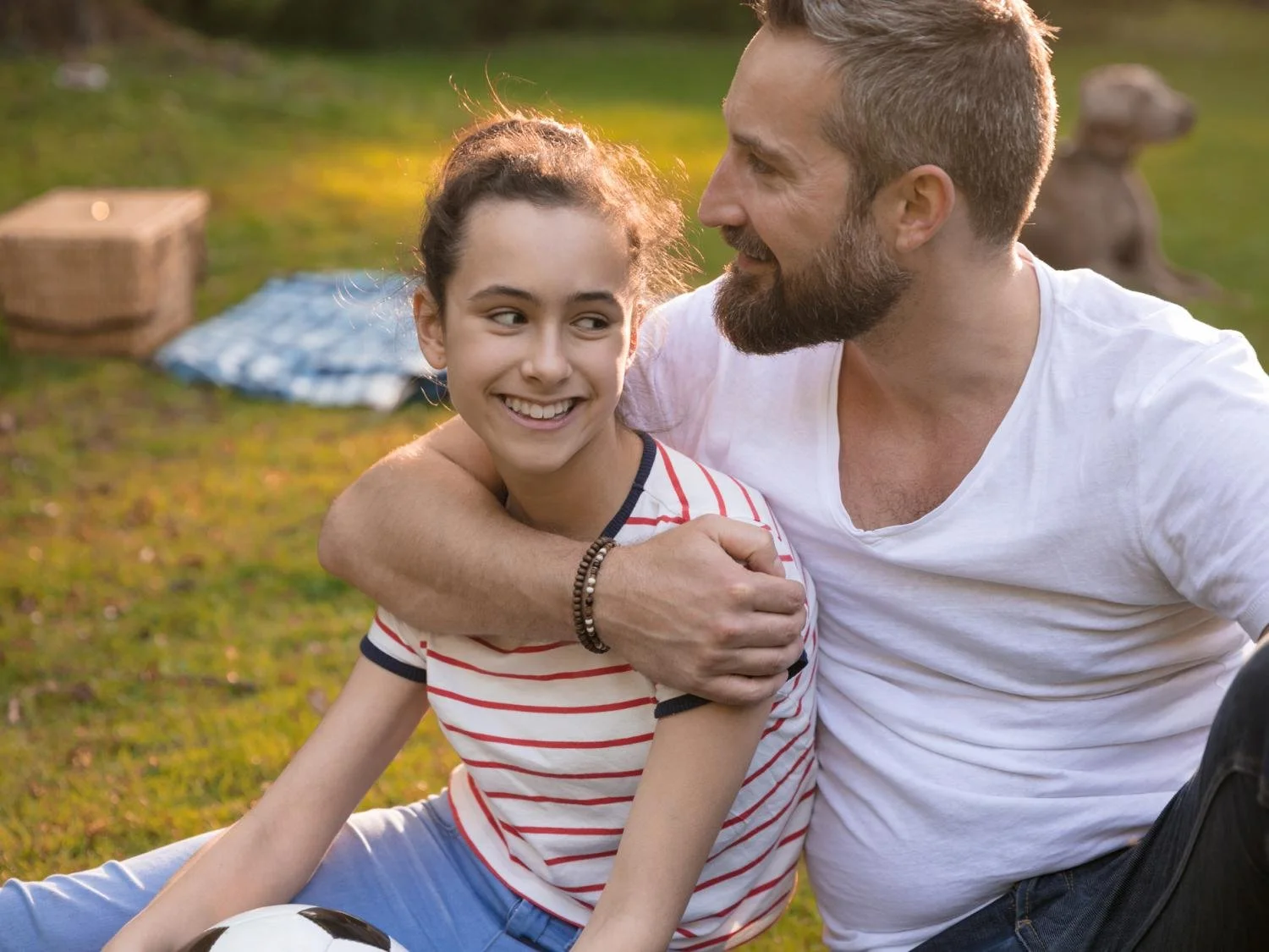 A man and a girl sitting on grass, smiling and embracing, outdoors during sunset, with a soccer ball nearby.