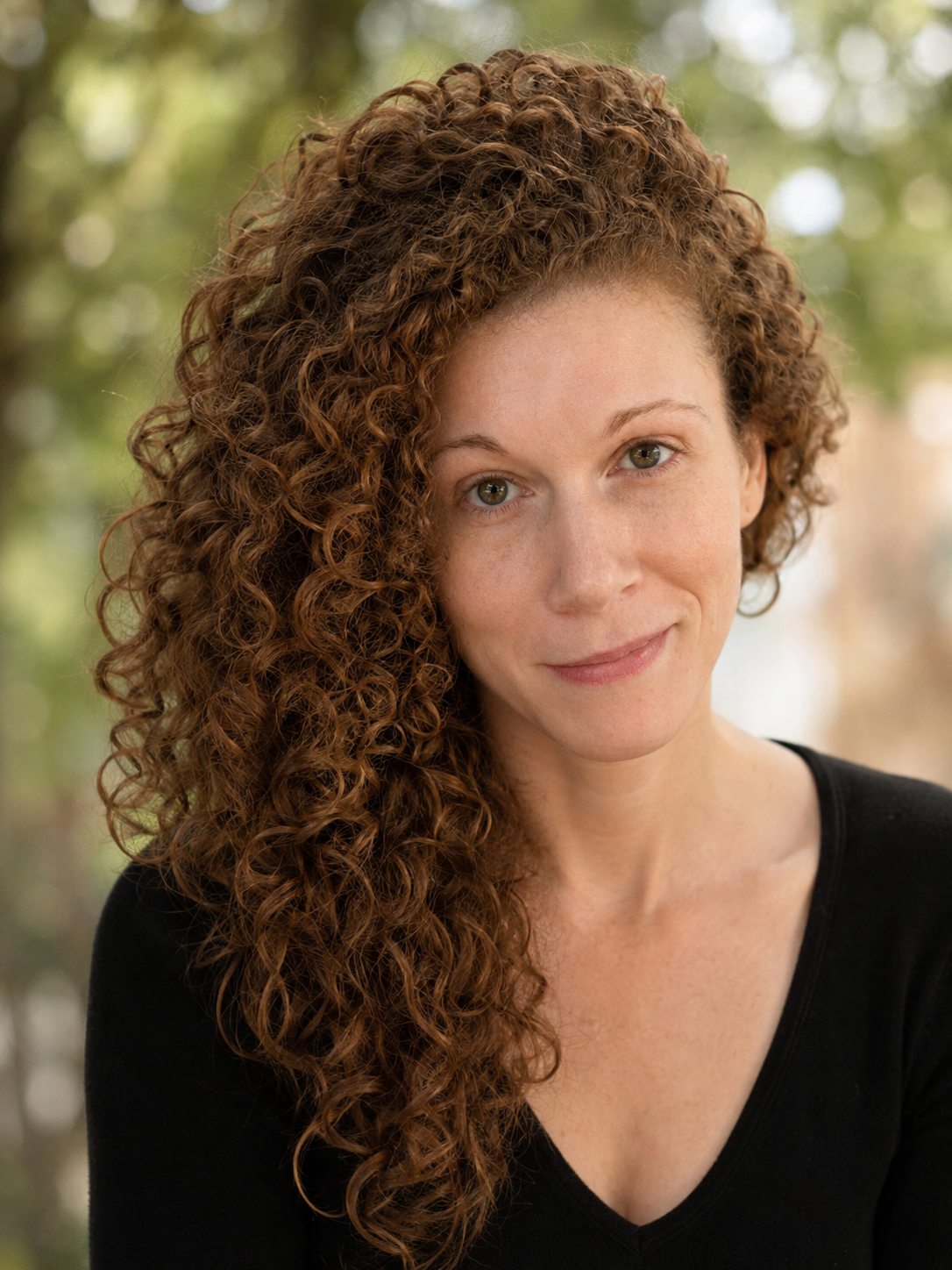 A woman with curly red hair, wearing a black top, smiling outdoors with blurred green background.