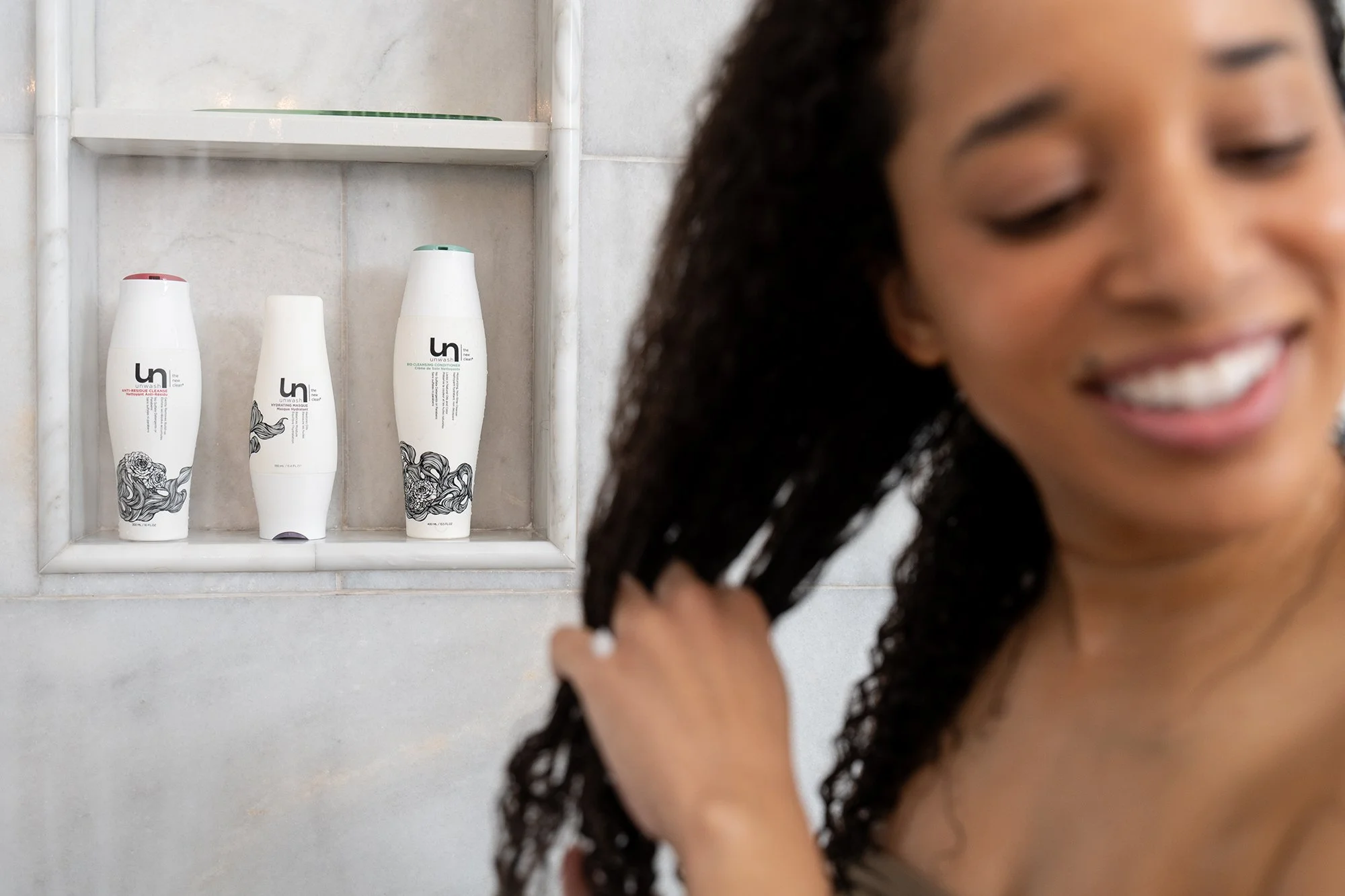 A woman with dark, curly hair smiling in a bathroom, with three Unwash products on a marble shelf behind her.