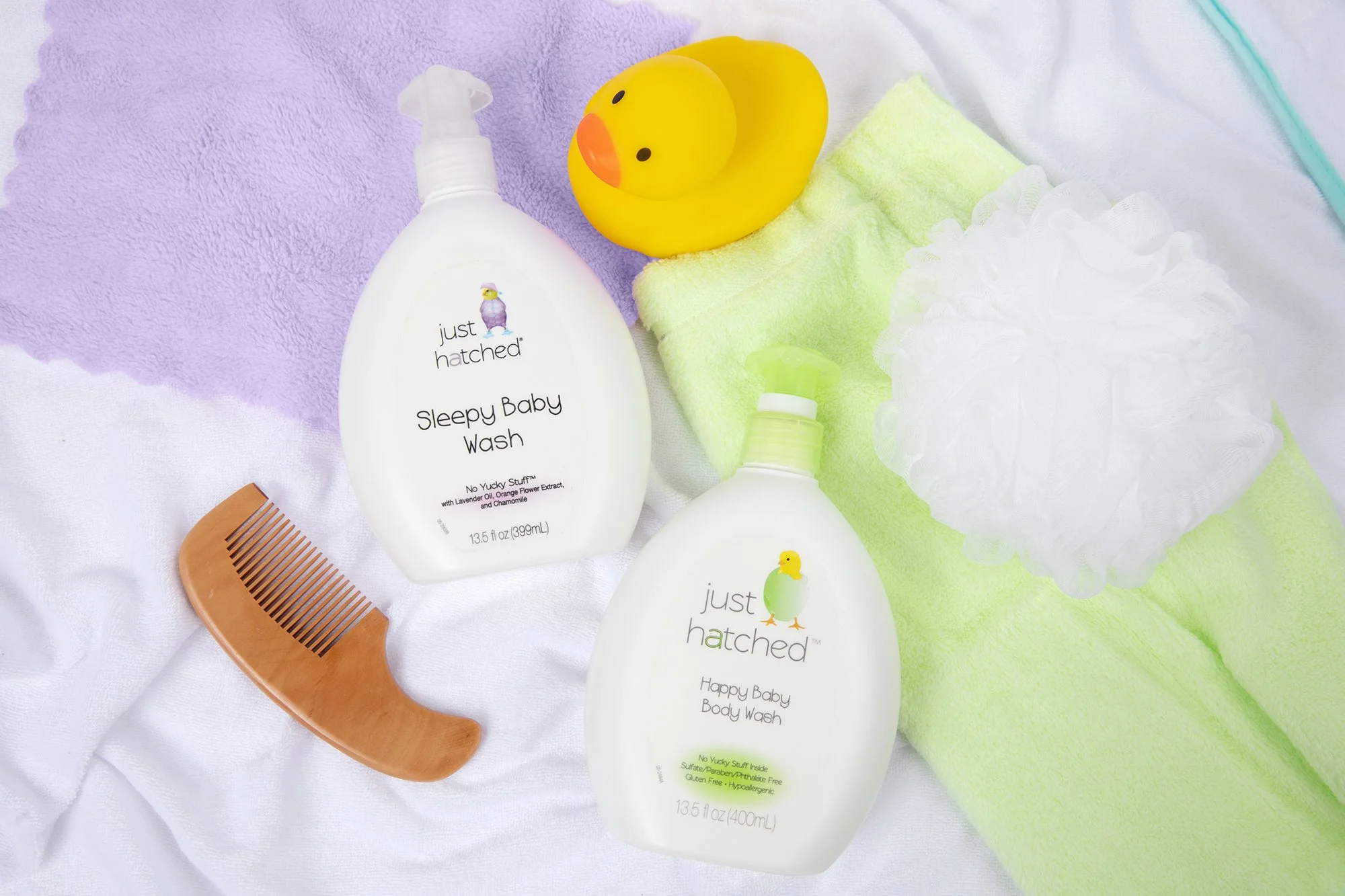 Baby bath supplies including two bottles of Just Hatched Sleepy Baby Wash, a yellow rubber duck, a wooden comb, a purple towel, and a white bath sponge, arranged on a white and pastel-colored towels.