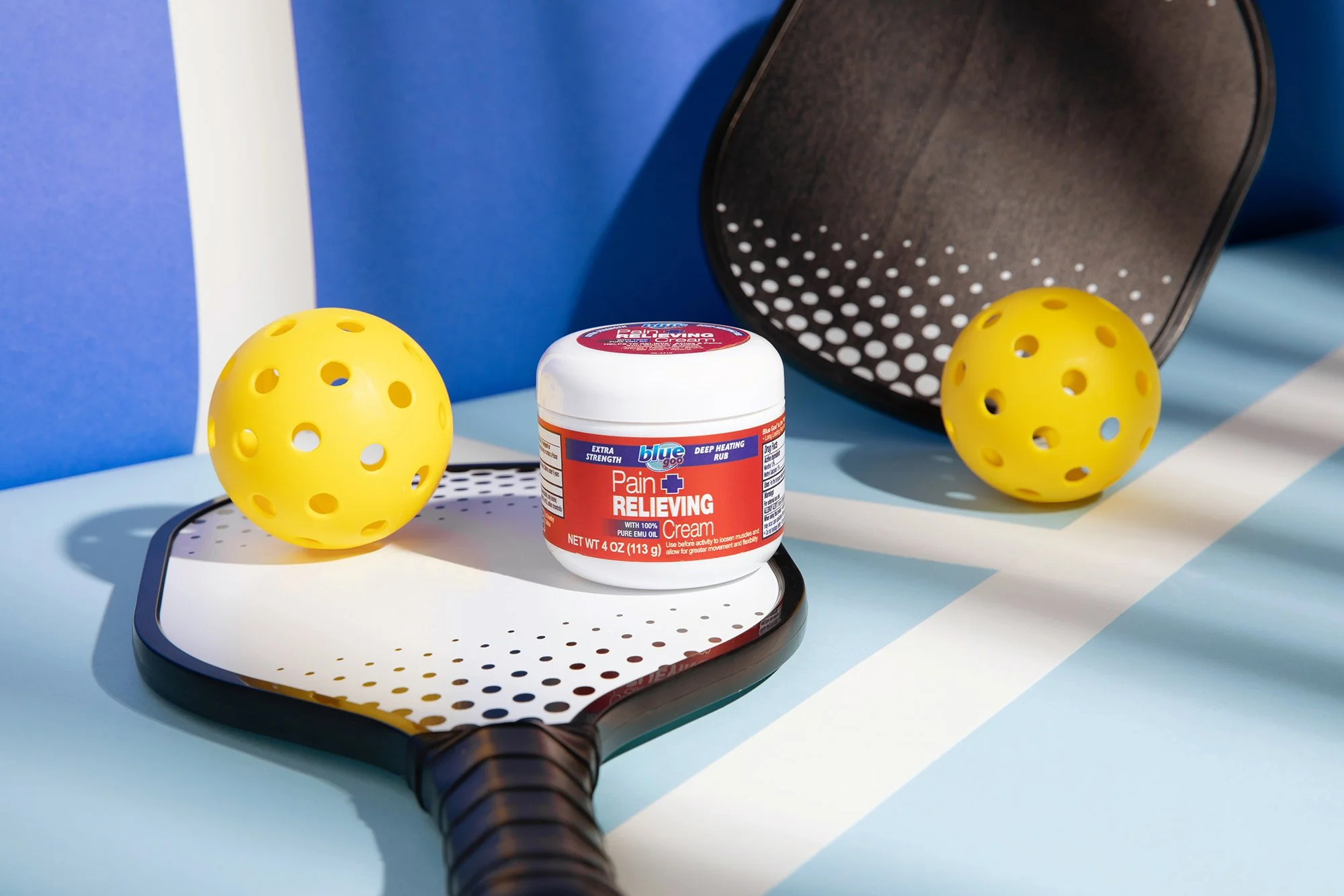 A container of pain relieving cream placed on a black and white tray, surrounded by yellow perforated balls, with a tray and a mirror in the background.