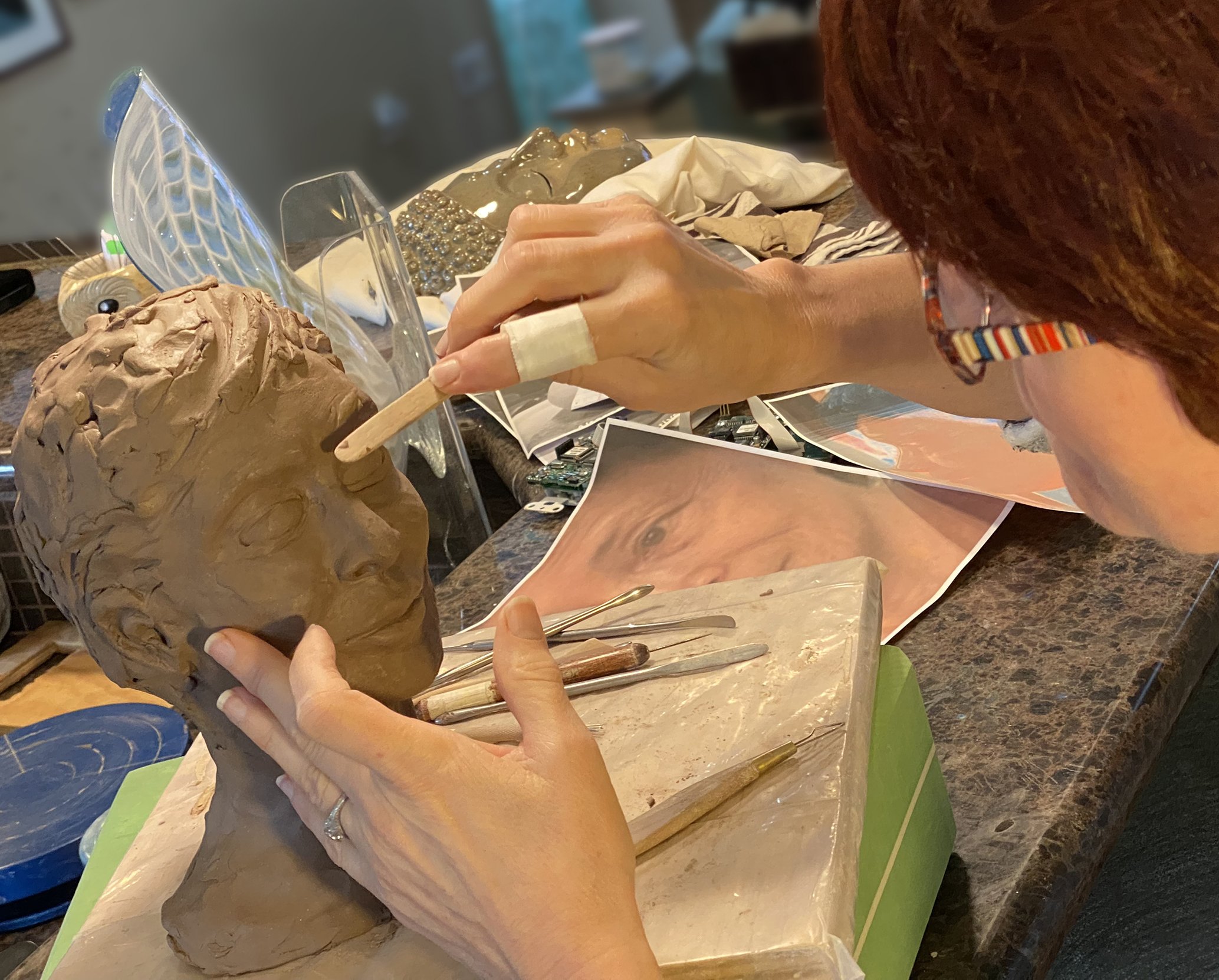 A person with red hair working on a clay sculpture at a worktable. The person's hands are visible, holding a tool and shaping a brown clay piece. There are various art supplies and photos spread out on the table, including a piece of paper, tools, and photographs of sculptures. The background is slightly blurred.