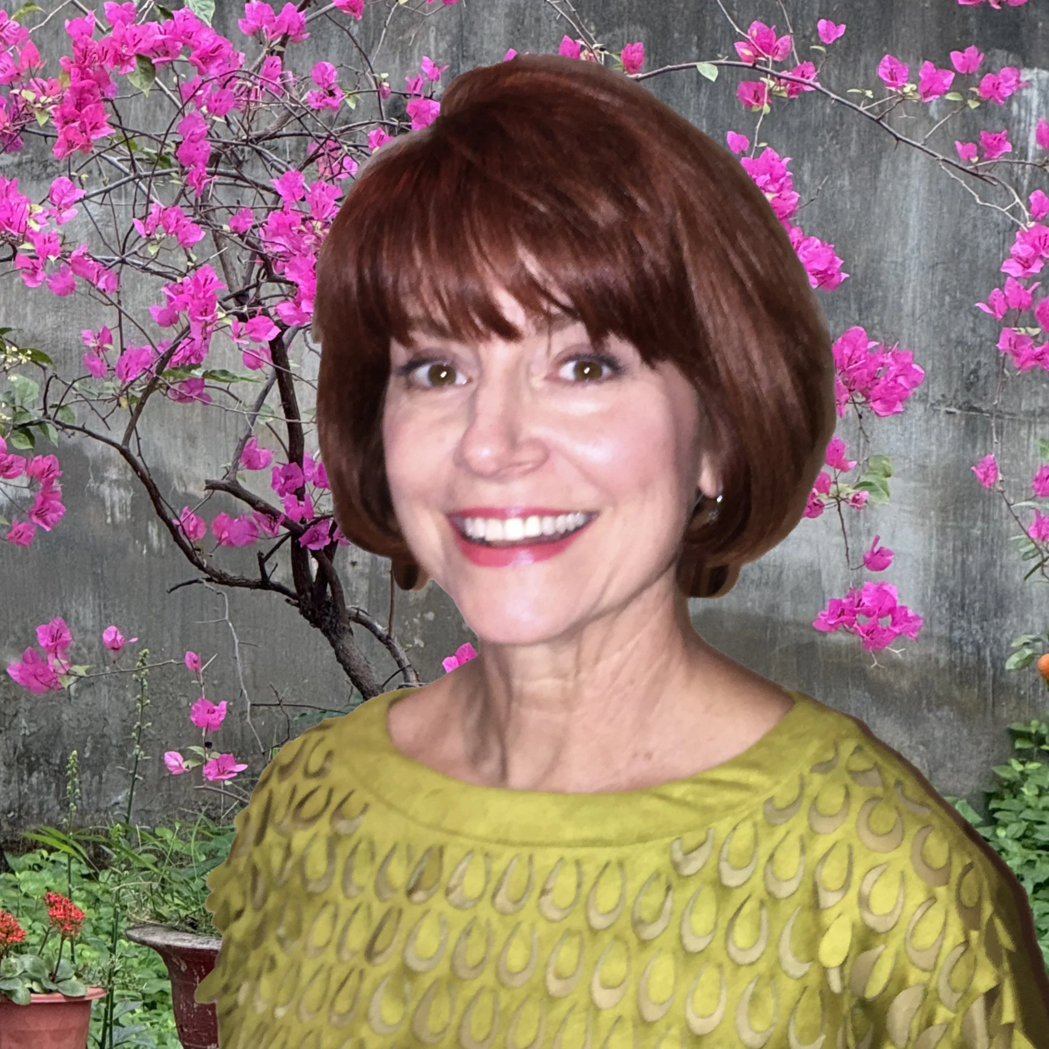 A woman with short brown hair, smiling, and wearing a green top, standing in front of a backdrop of pink bougainvillea flowers and green foliage.