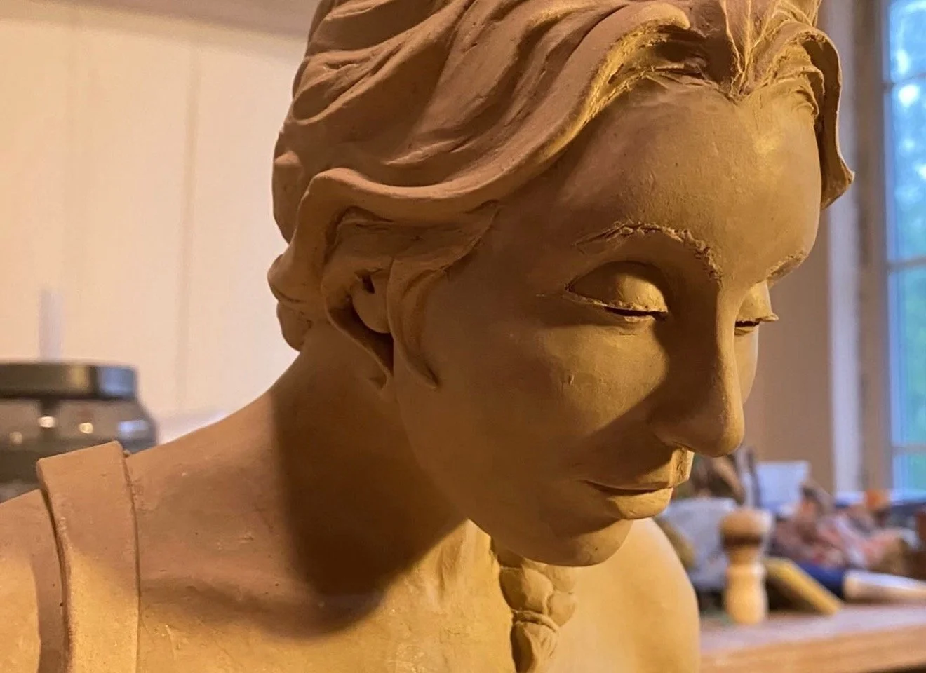 A detailed wooden sculpture of a person's head and shoulders with closed eyes, long wavy hair, and a gentle expression, set against a background of a room with a window and various objects.