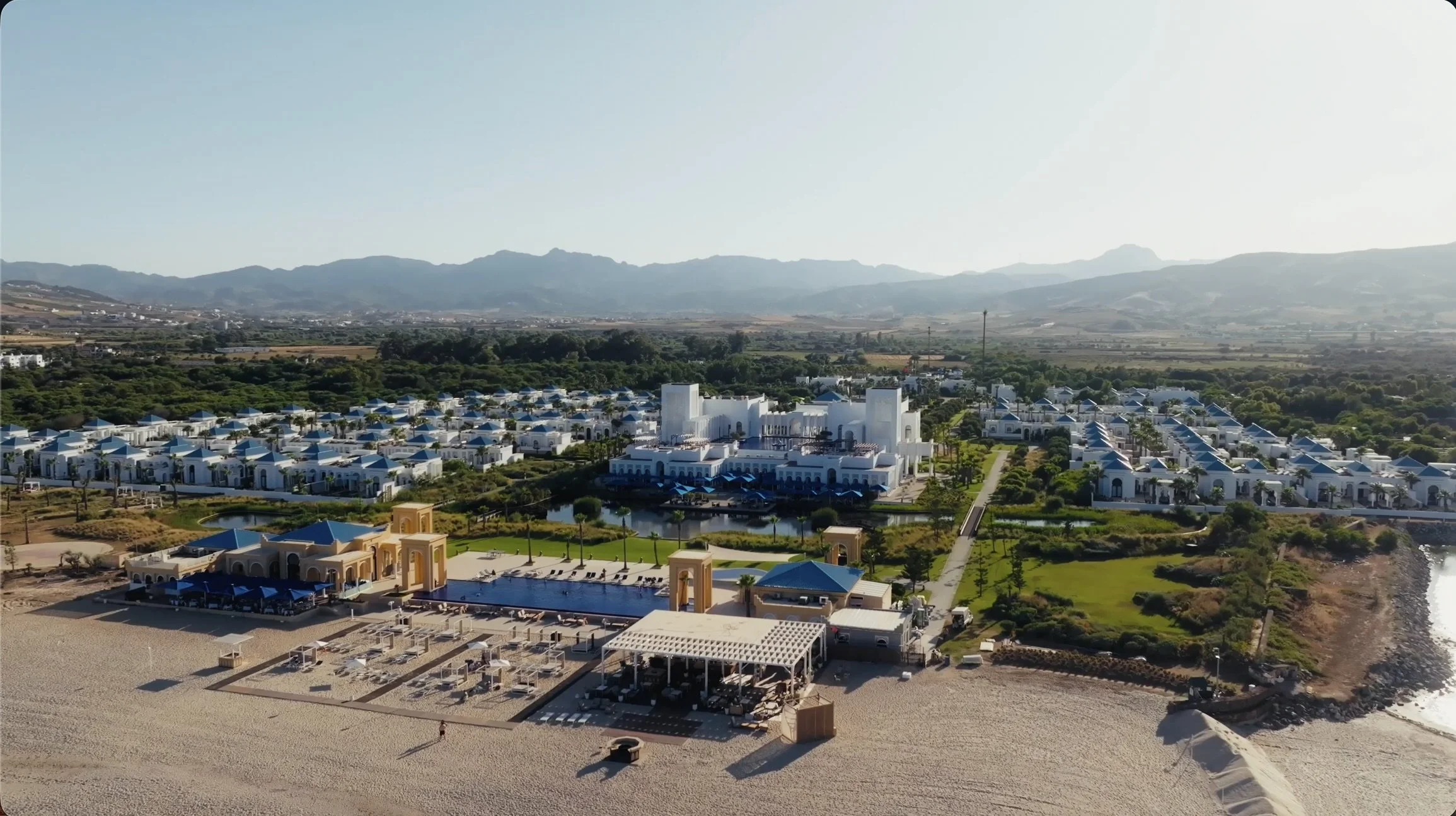 Aerial view of a resort with white buildings and blue roofs, surrounded by greenery and mountains in the background.