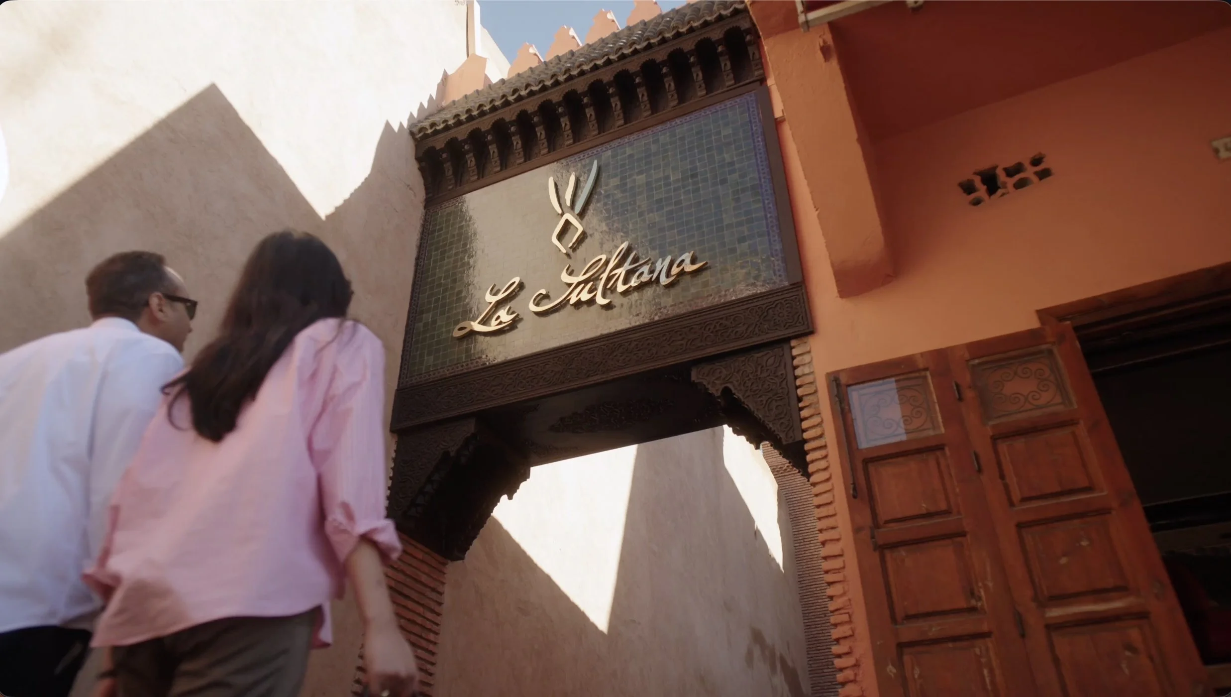 A man and a woman walking past a decorative archway with the sign 'La Sultana' and traditional Moroccan architectural details.