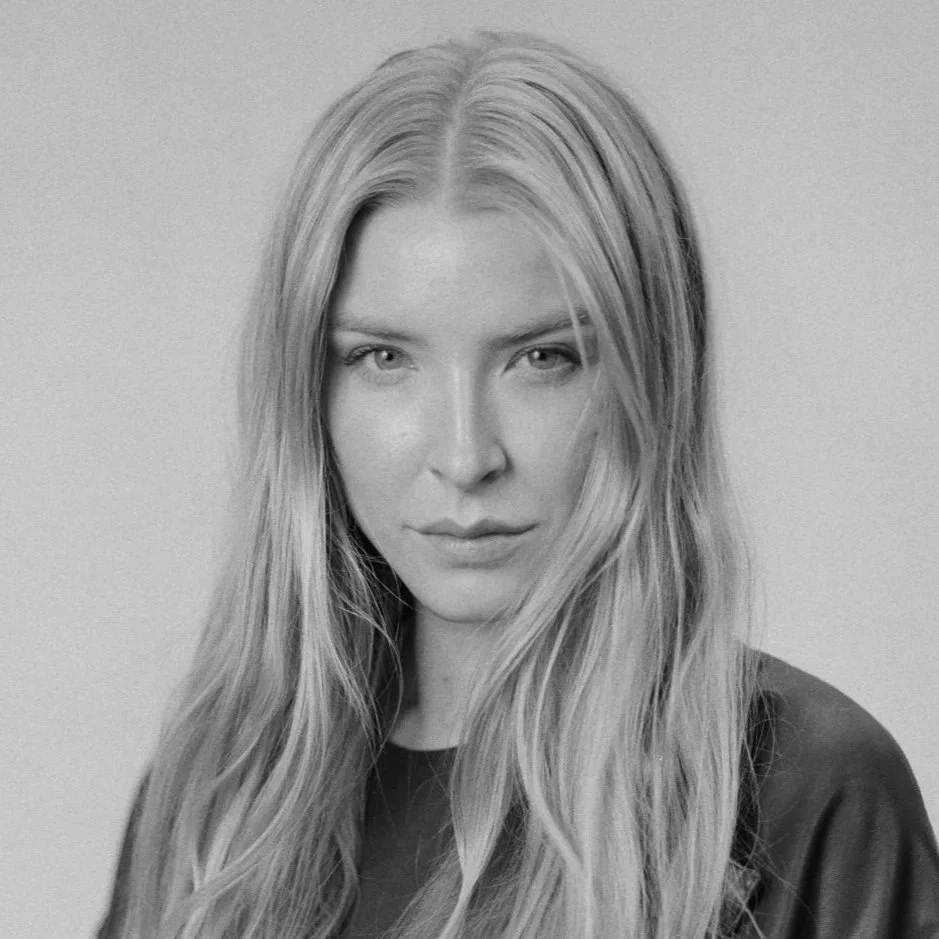 A black and white portrait of a woman with long, wavy hair wearing a leather jacket, looking directly at the camera.