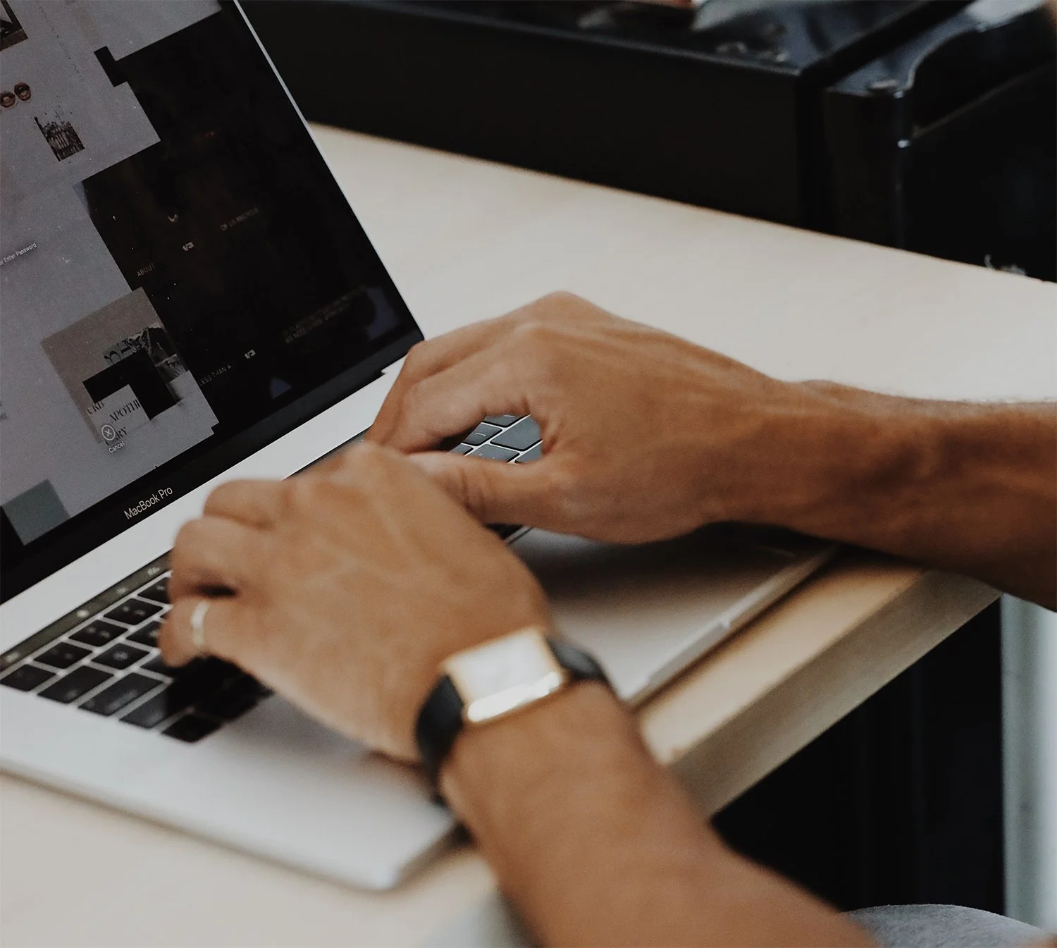 Person using a MacBook Pro laptop at a desk, with their left hand on the keyboard and wearing a black watch with a gold face.
