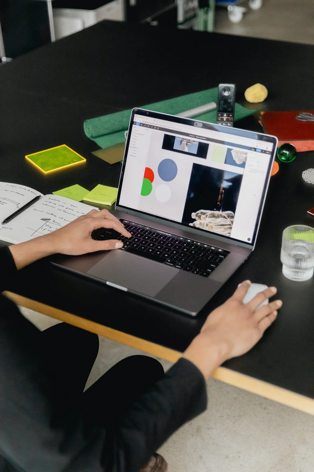 A person working on a laptop at a black table with various papers, sticky notes, a glass of water, and colorful objects.