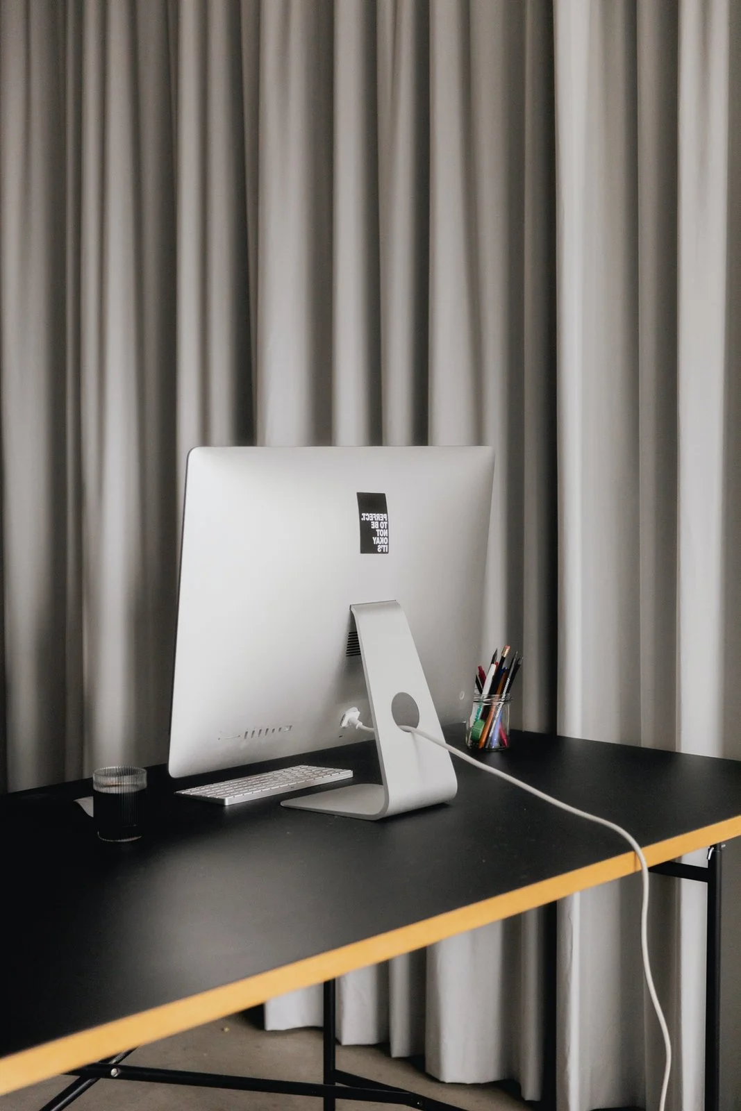 An iMac computer on a black desk with a white keyboard, a glass of dark liquid, and a container of pens in front of a gray curtain.