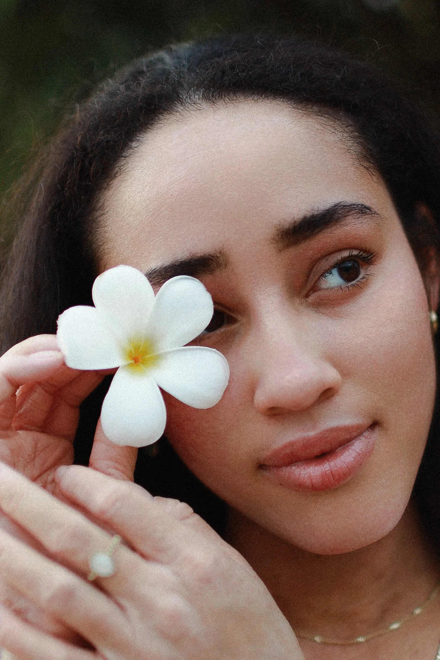 A woman holding a white flower near her face, with a serene expression and natural makeup.