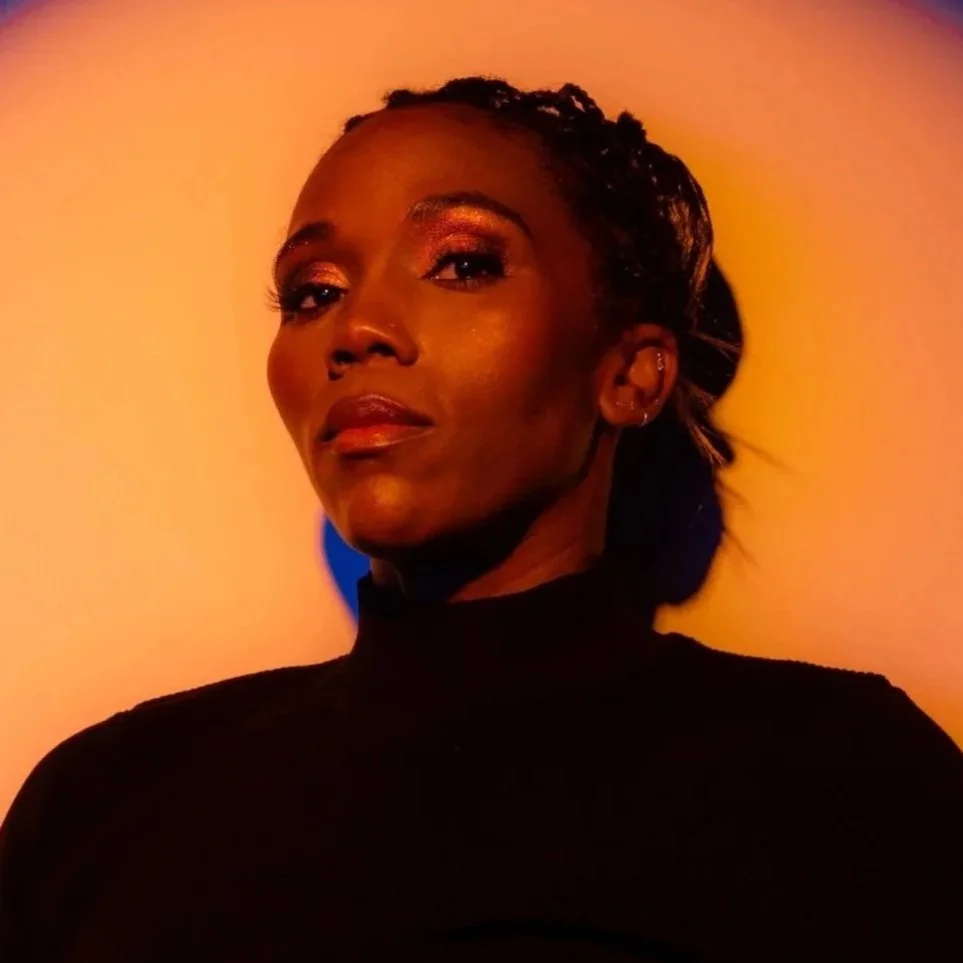 A woman with dark skin and short curly hair posing in front of a colorful, circular light display with a blue background.