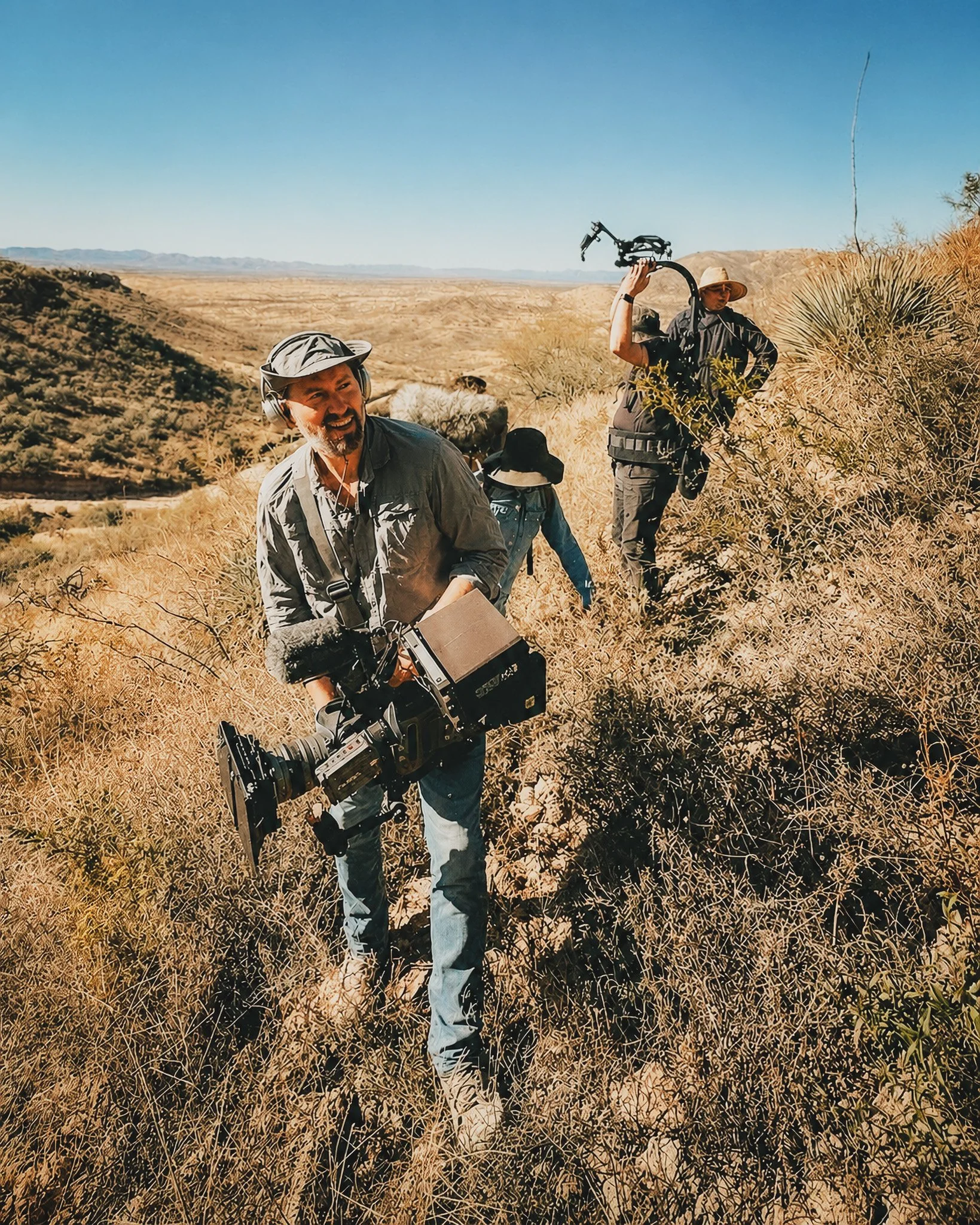 Three men in outdoor gear in a desert landscape with yellow grass, shrubs, hills, and a clear blue sky.