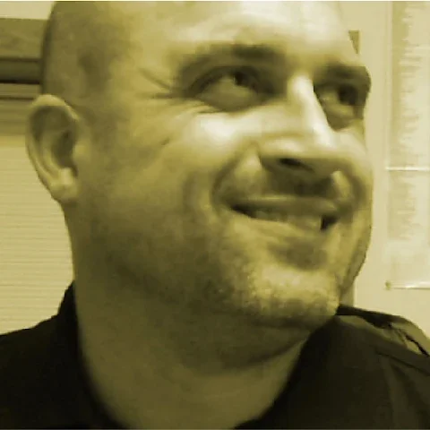 A smiling man in a dark uniform with a shaved head, looking to the right. The background includes a window with closed blinds.
