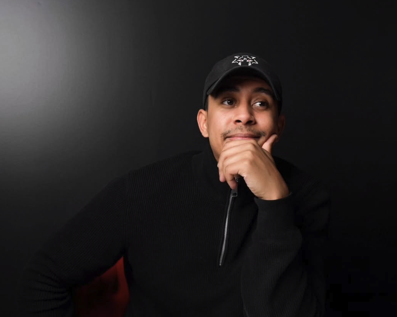 A young man wearing a black cap and black zip-up sweater sitting against a black background, resting his chin on his hand and looking thoughtfully to the side.