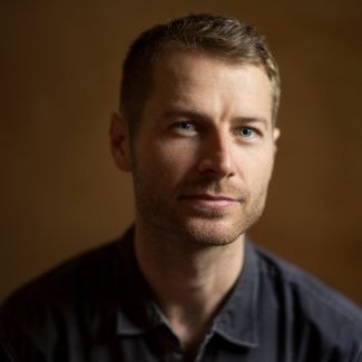 Portrait of a man with short hair and a slight smile, wearing a dark shirt against a warm background.