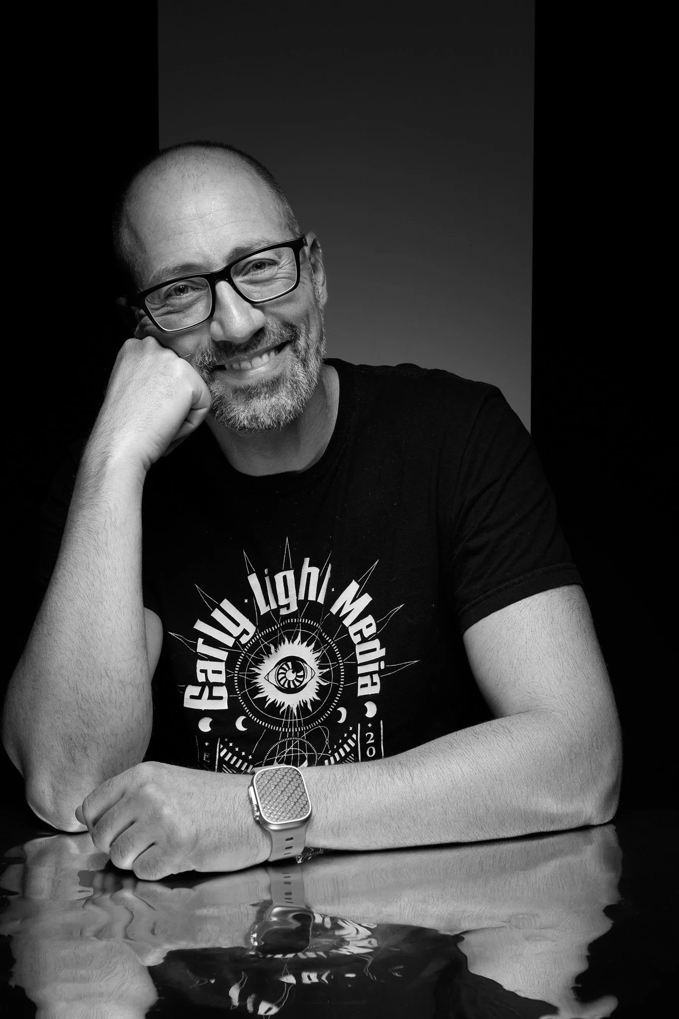 Darren Durlach - A man with glasses and a beard, smiling and resting his head on his hand, sitting at a reflective surface. He is wearing a graphic T-shirt and a wristwatch.
