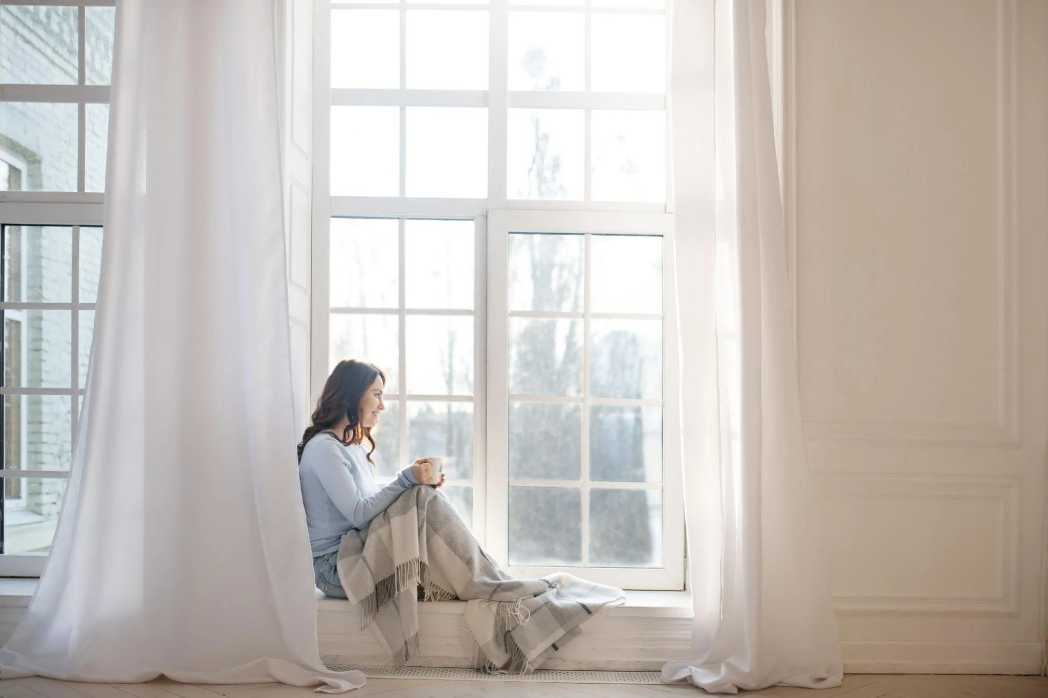 A young woman sitting by a bright window wrapped in a cozy blanket, representing self-compassion, emotional healing, and personal well-being.