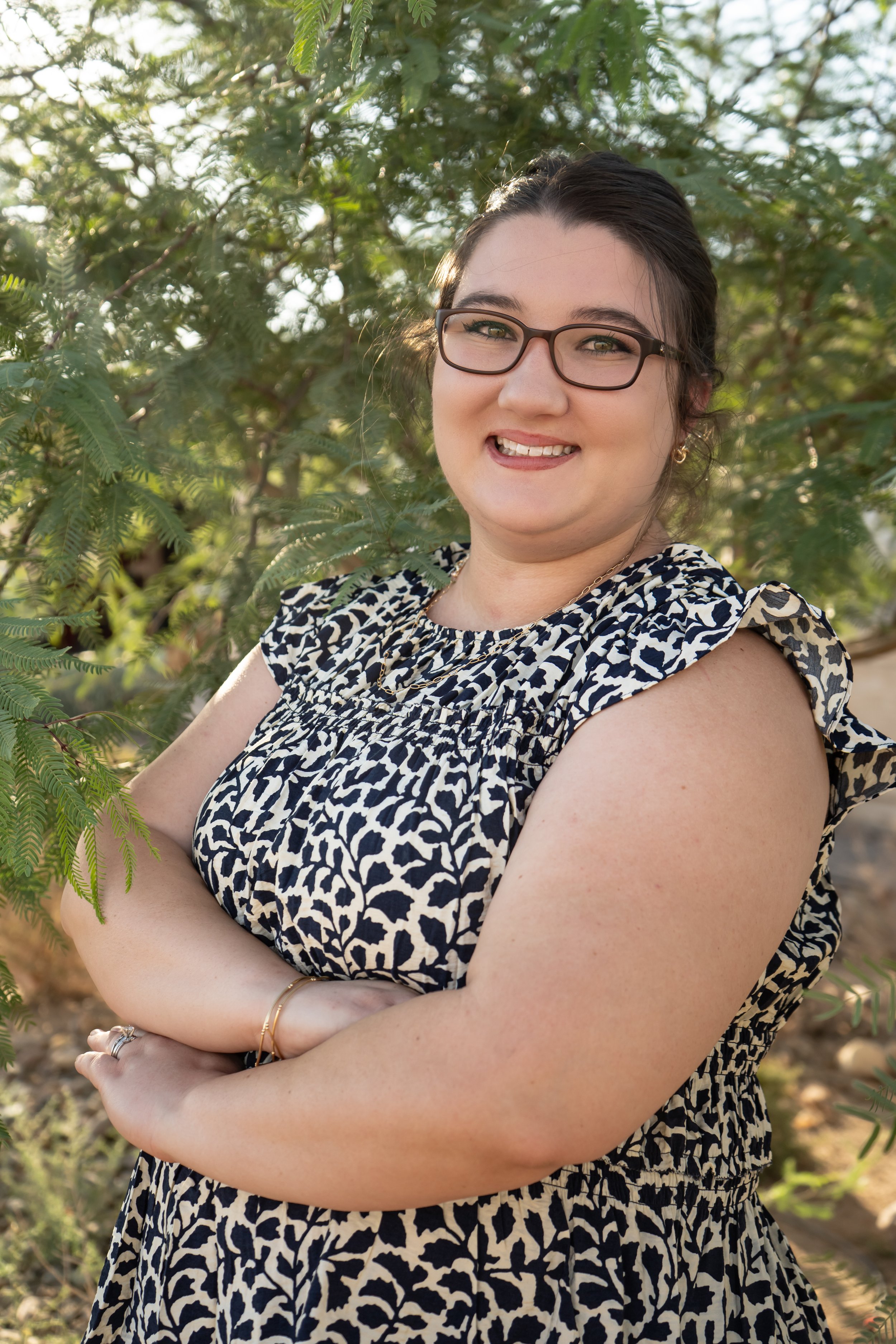 A woman with glasses and dark hair in an updo, smiling with her arms crossed, standing outdoors among green foliage during daytime.