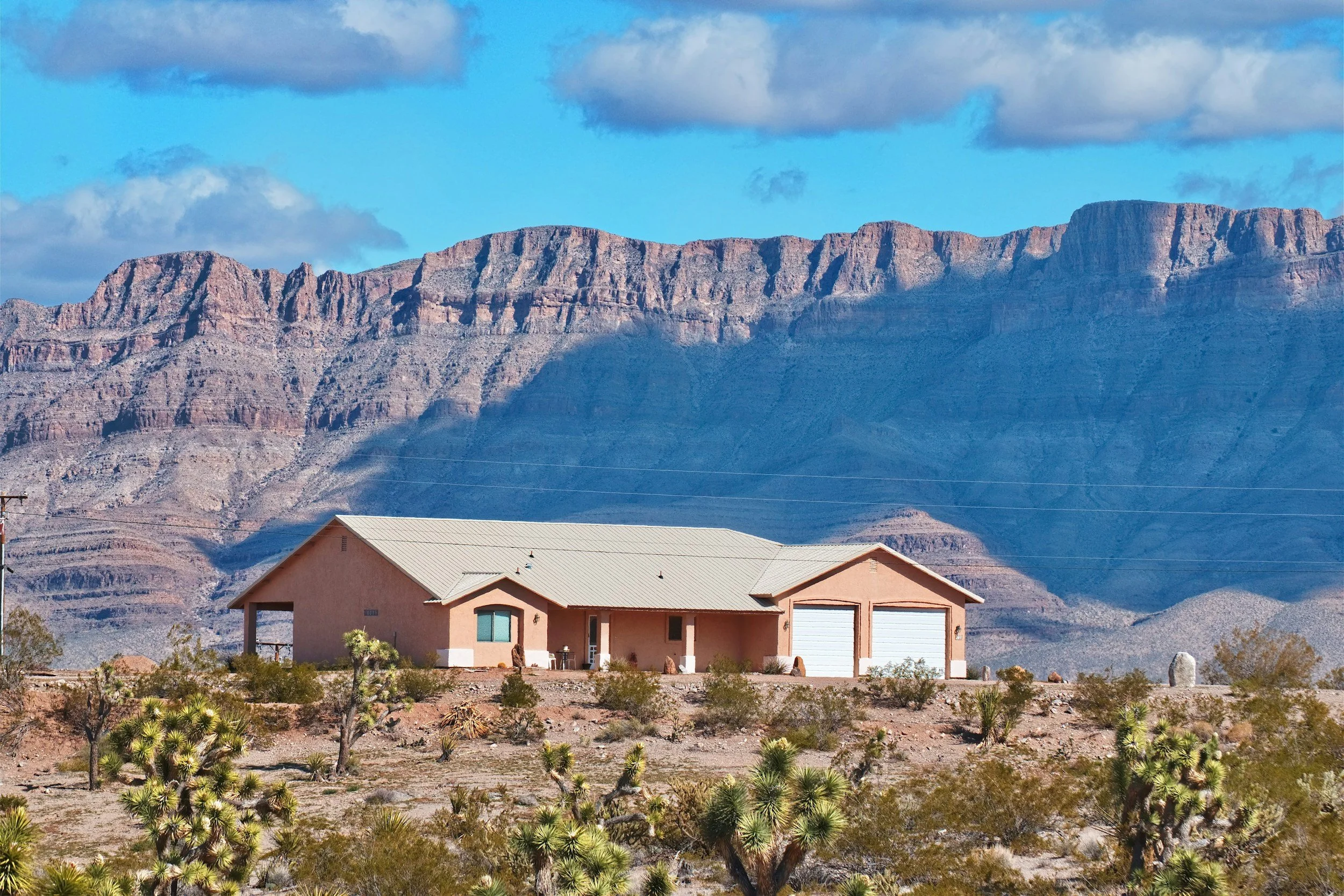 A house in a desert landscape with mountains in the background and a bright blue sky with clouds.