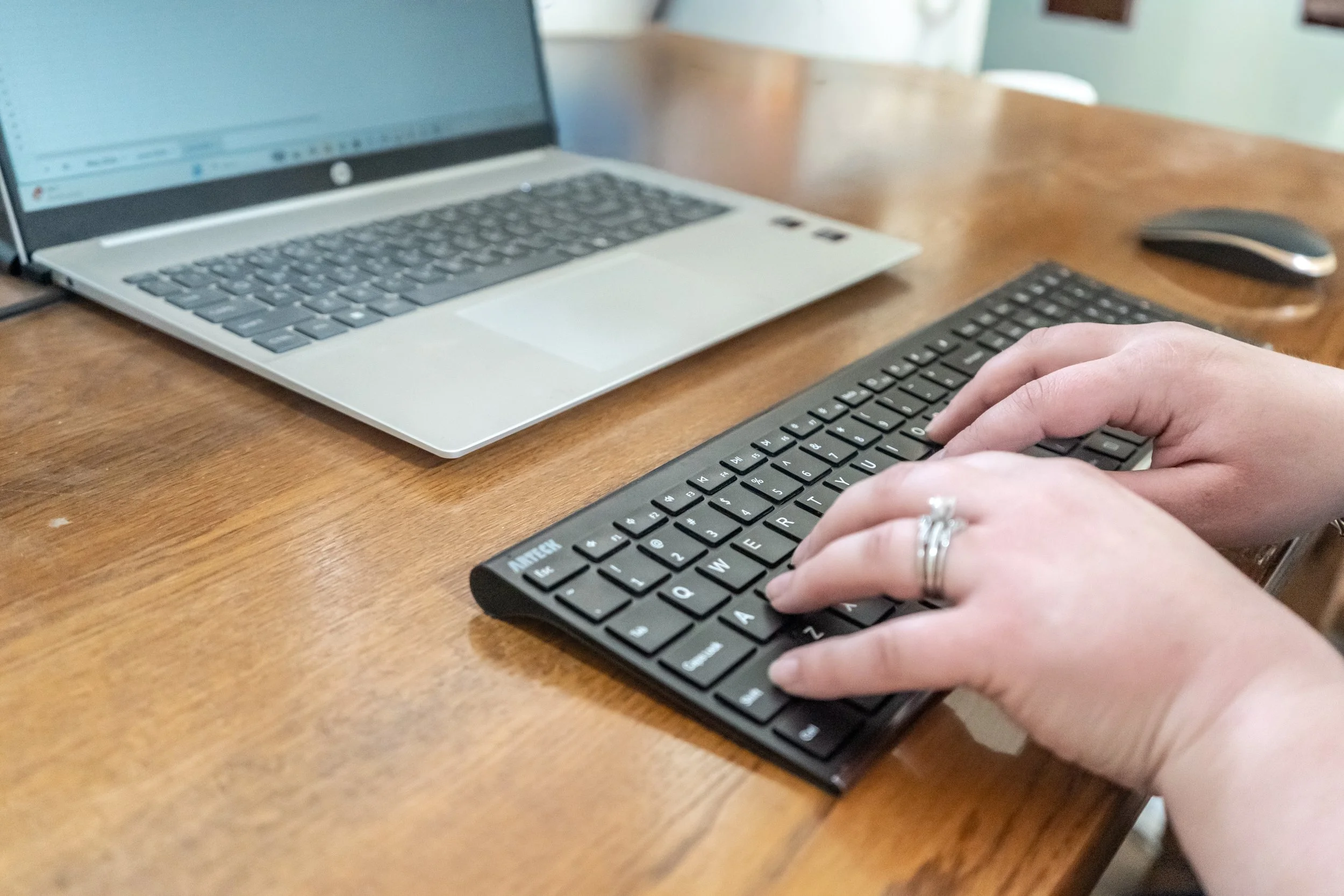 Person typing on a black wireless keyboard at a wooden desk with a silver HP laptop and a black wireless mouse.