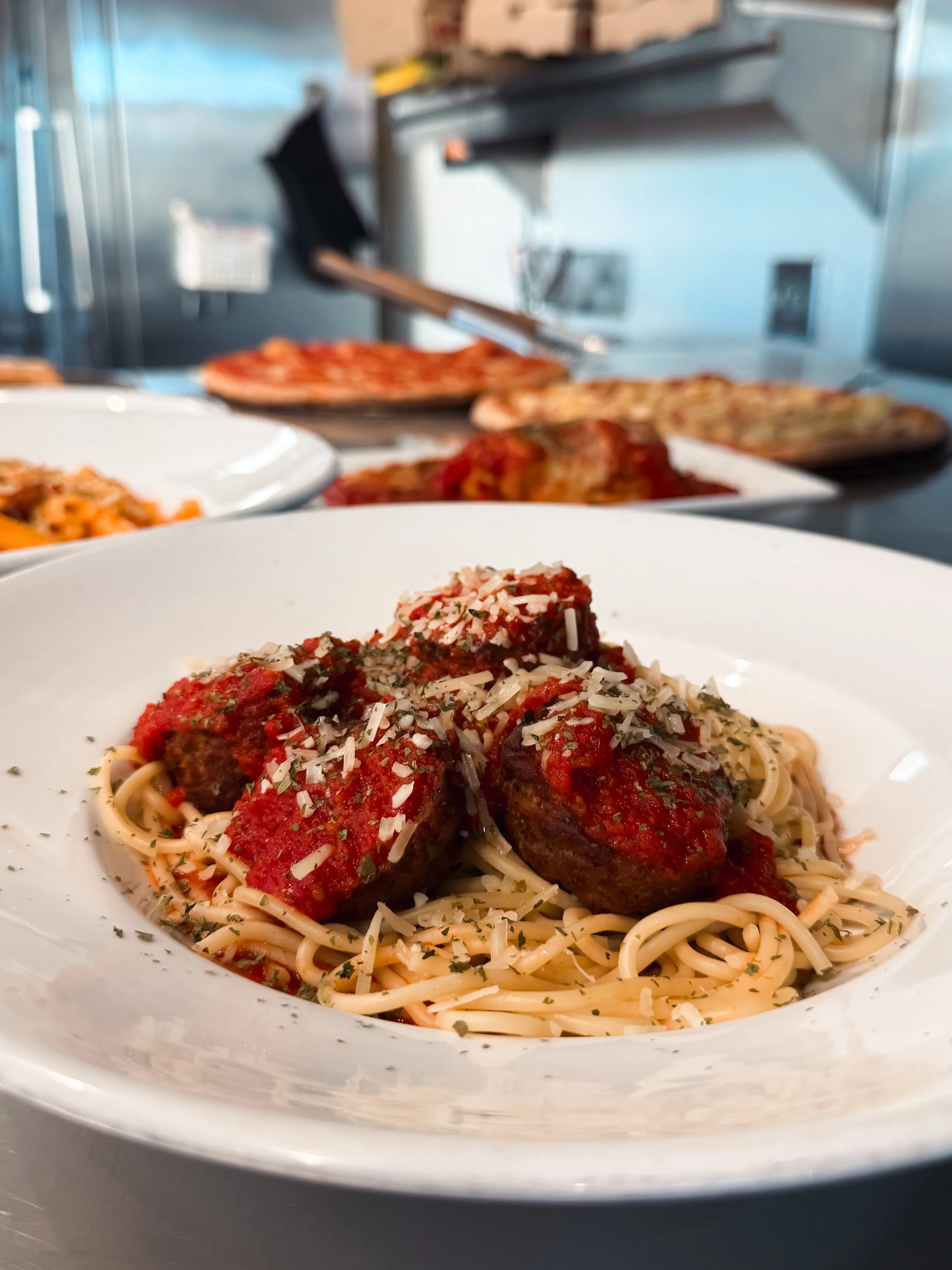 A plate of spaghetti with marinara sauce topped with meatballs and sprinkled with grated cheese and herbs, in the foreground. In the background, there are various pizzas on display in a restaurant or cafeteria setting.