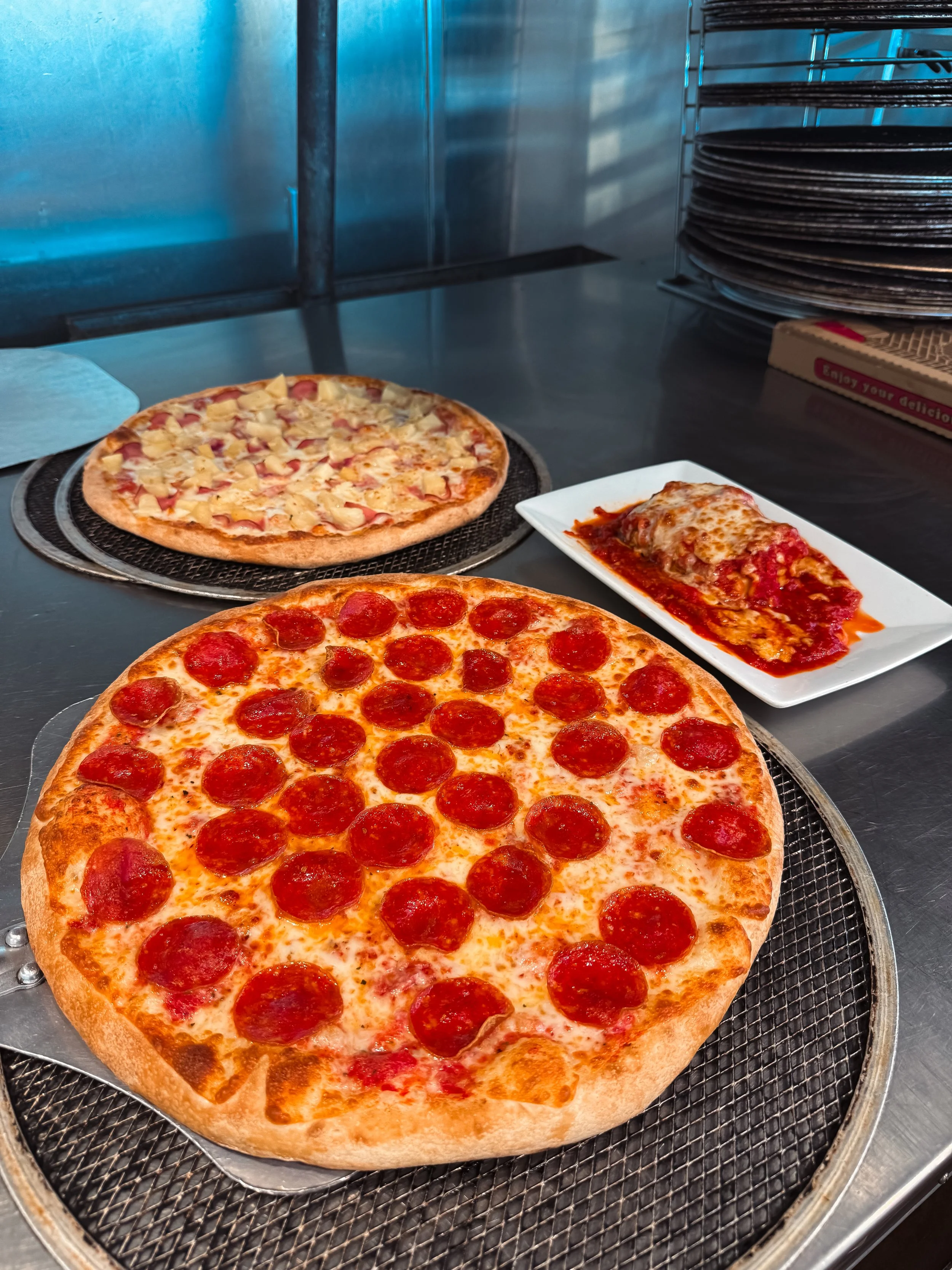 Close-up of a pepperoni pizza on a wire rack, with another pizza and a plate of lasagna in the background.