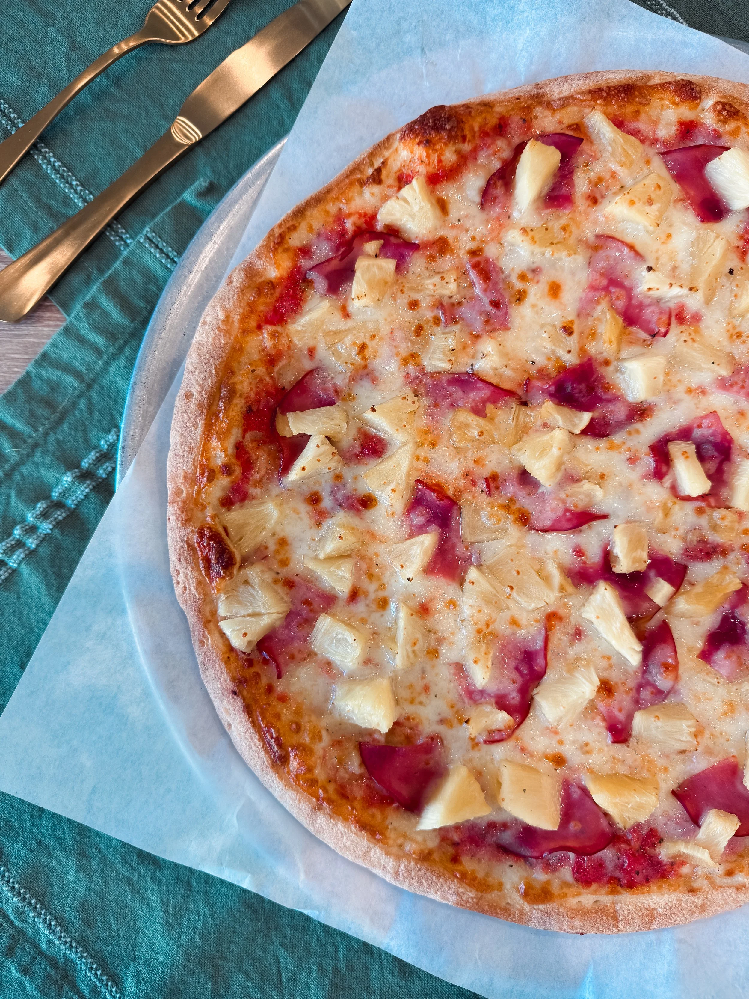 Close-up of a pineapple and ham pizza on a piece of parchment paper, with a green napkin and a fork and knife beside it.