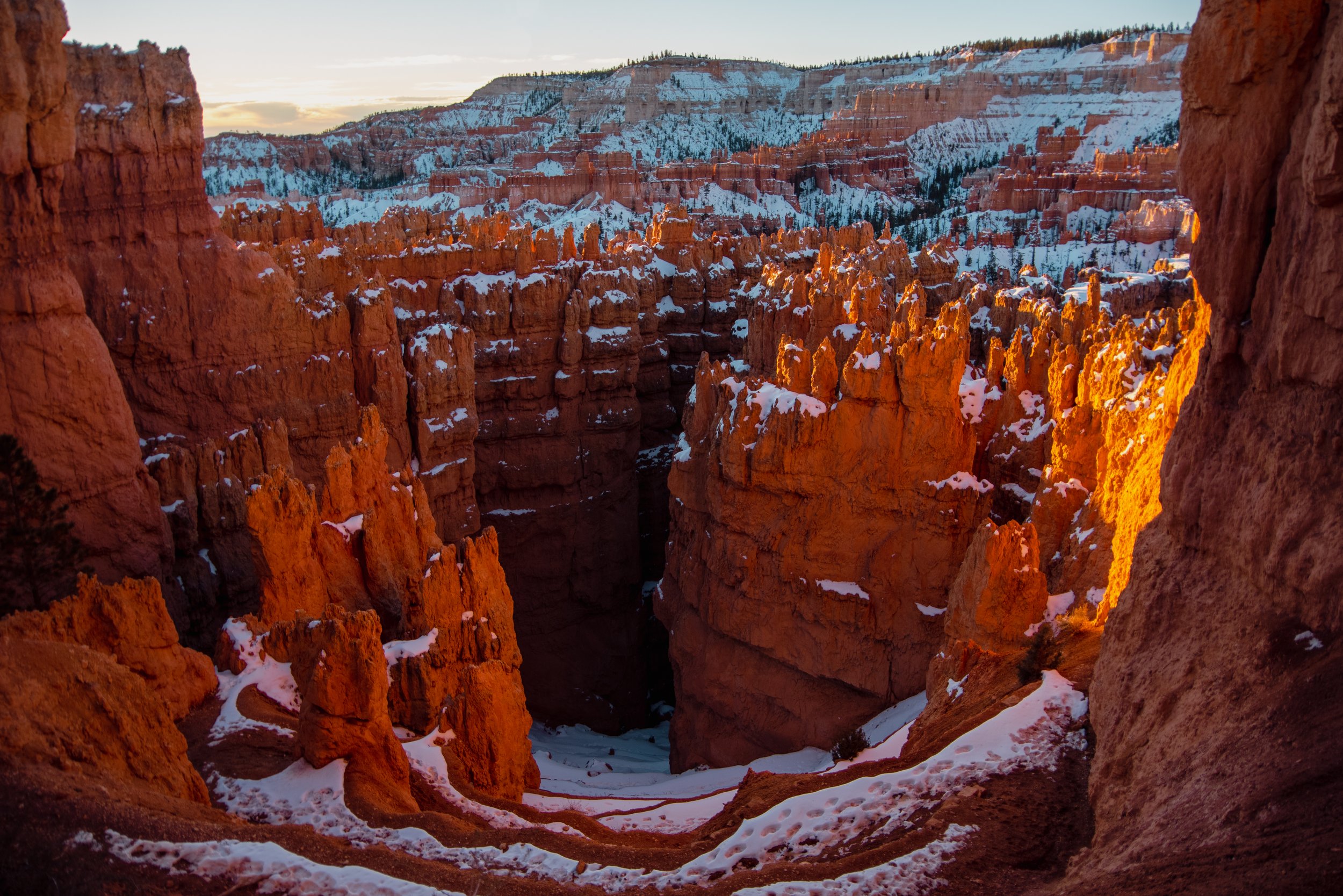 Sunset over ancient red rock formations in a canyon with snow-covered peaks in the background.