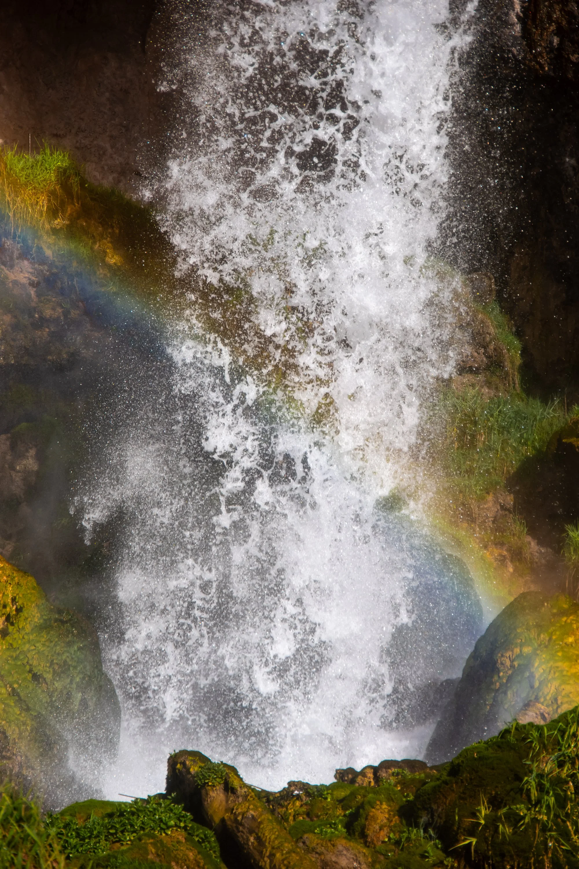 A waterfall cascading down rocks surrounded by green moss and plants, with a rainbow visible at the bottom left.
